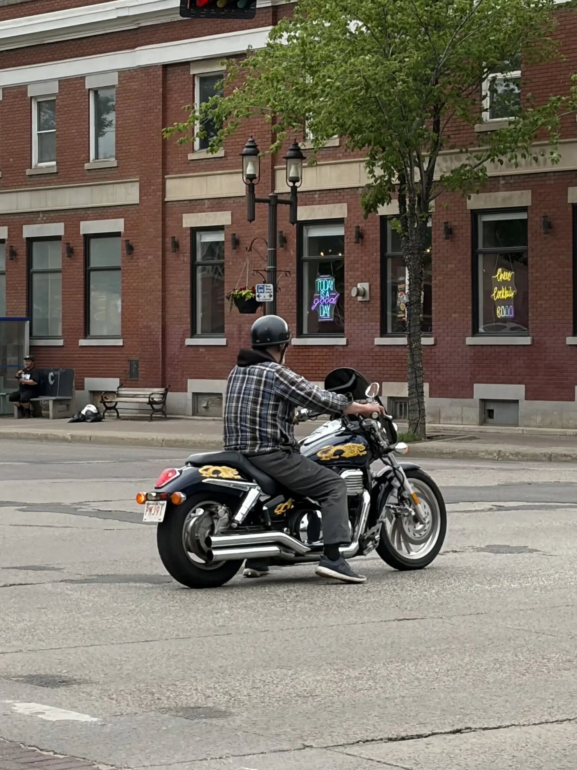 A man wearing a black helmet and jacket sits on a dark motorcycle at a city street intersection.