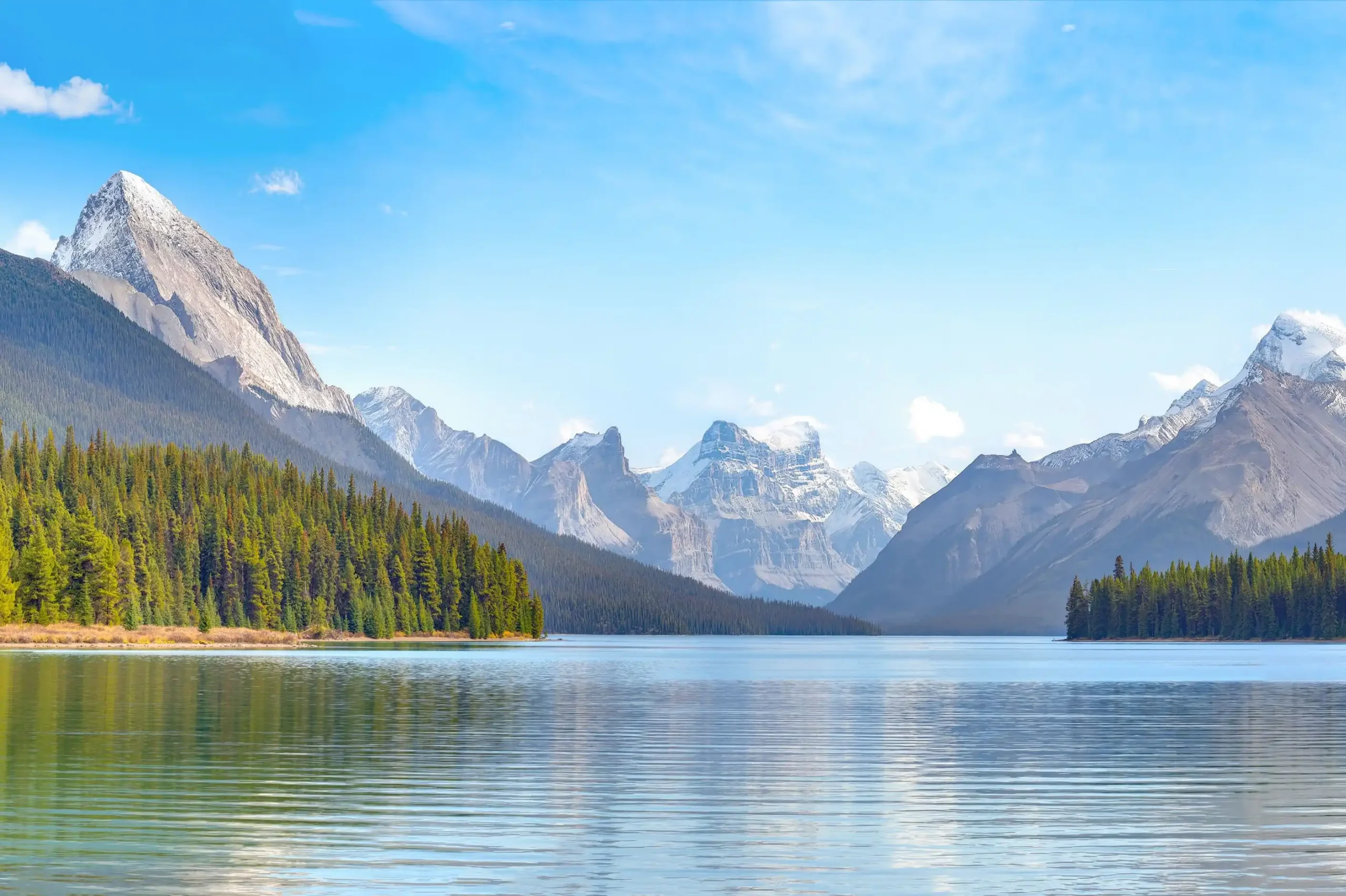 A calm mountain lake surrounded by evergreen forest, with snow-capped peaks reflected in the water under a blue sky.