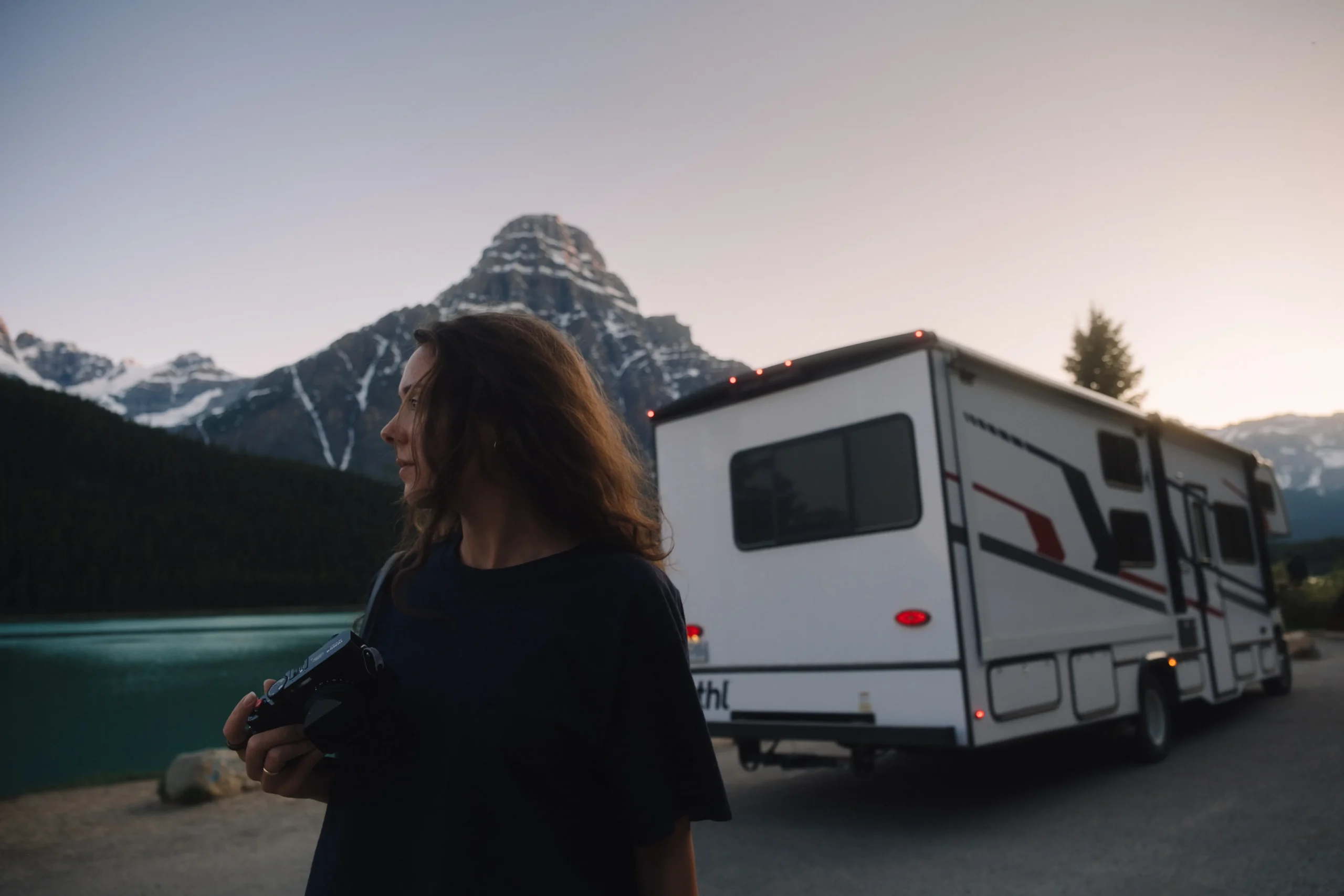 A woman holding a camera stands in the foreground, looking toward a large white motorhome parked beside a turquoise lake. In the background, a jagged, snow-dusted mountain peak rises under a clear twilight sky.