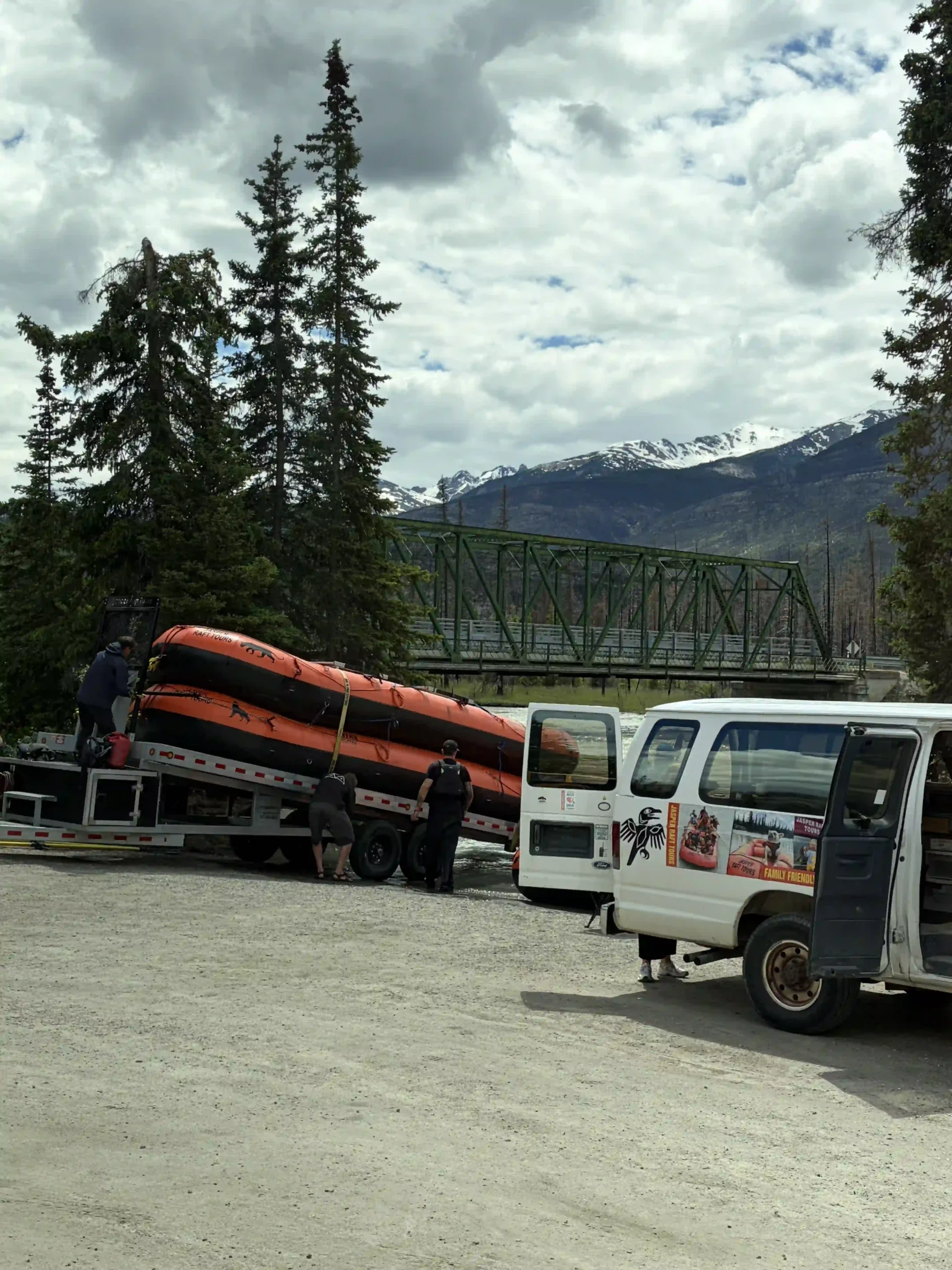 Large orange and black inflatable river rafts are stacked on a flatbed trailer hooked to a white passenger van with its doors open. Crew members are adjusting the gear in a gravel lot.