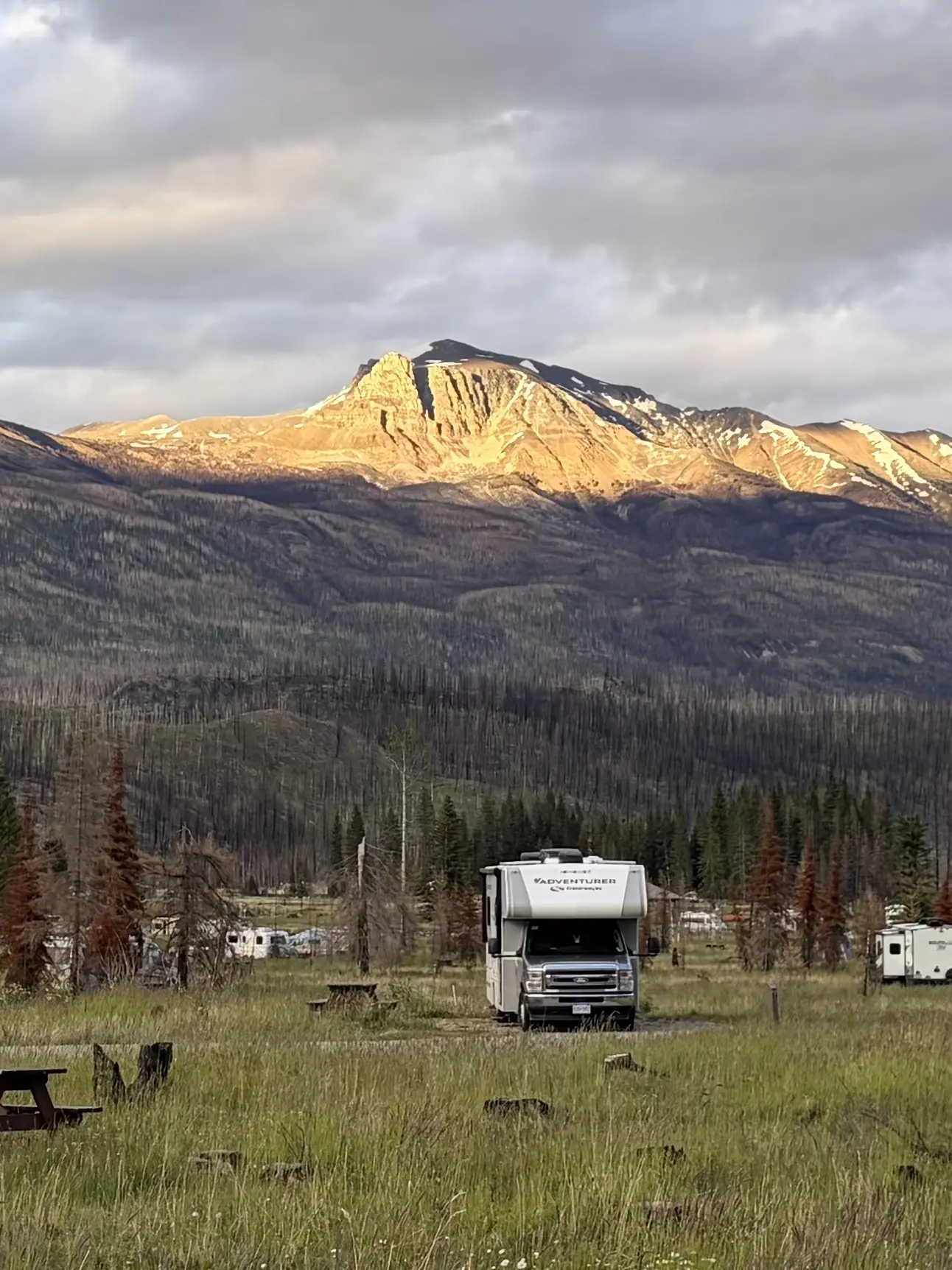 A white class C motorhome RV is parked in a grassy campsite slot at Whistlers Campground. In the background, a massive mountain peak is brightly illuminated by glowing golden sunlight under a dramatic, cloudy sky.