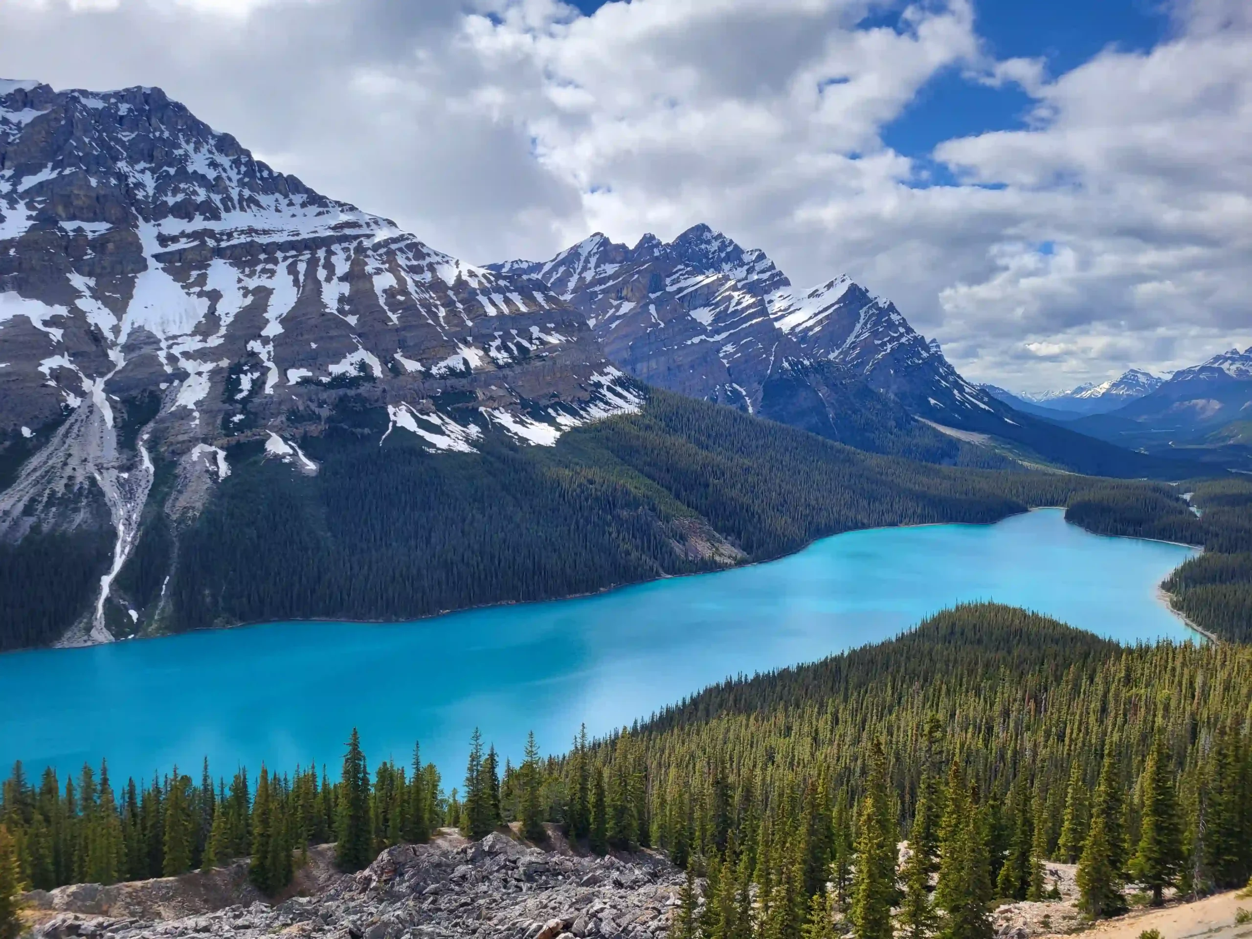 The lake is surrounded by dense green pine forests, with massive, rocky mountain peaks featuring patches of white snow.