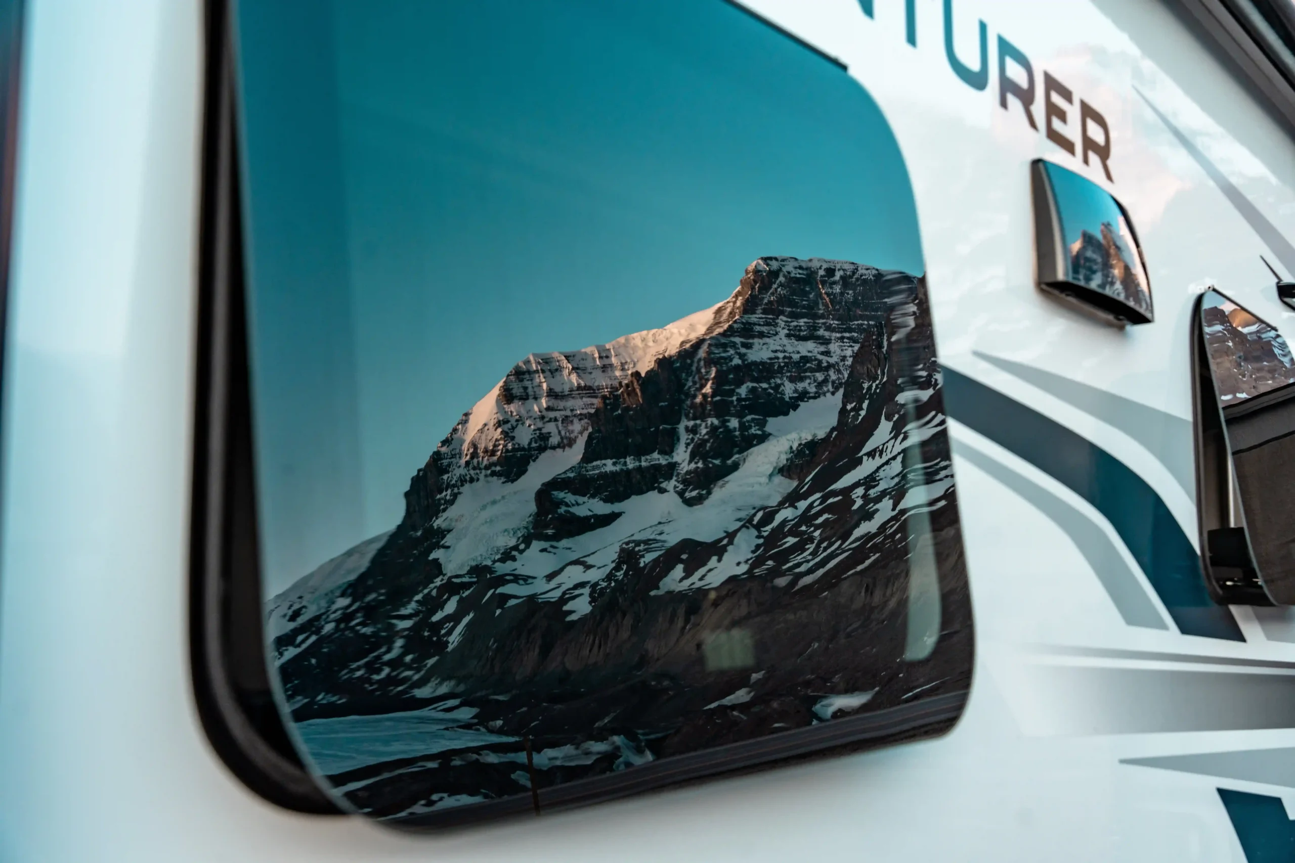 A close-up, artistic shot of a snow-capped mountain peak reflected perfectly in the dark-tinted window of a white motorhome.