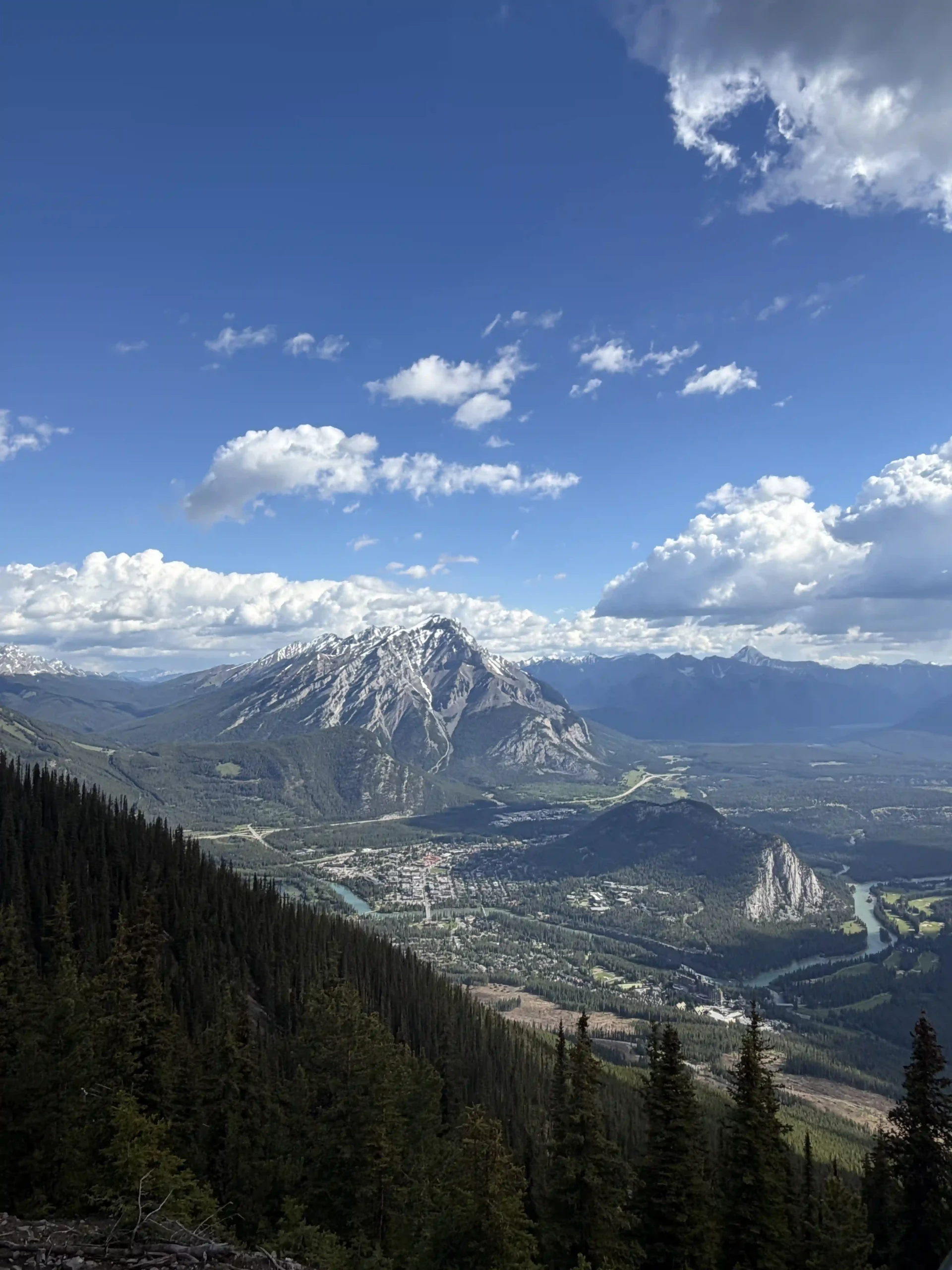Panoramic view of the town of Banff nestled in a mountain valley, with a winding river and a massive snow-capped peak under a bright blue sky with fluffy white clouds.