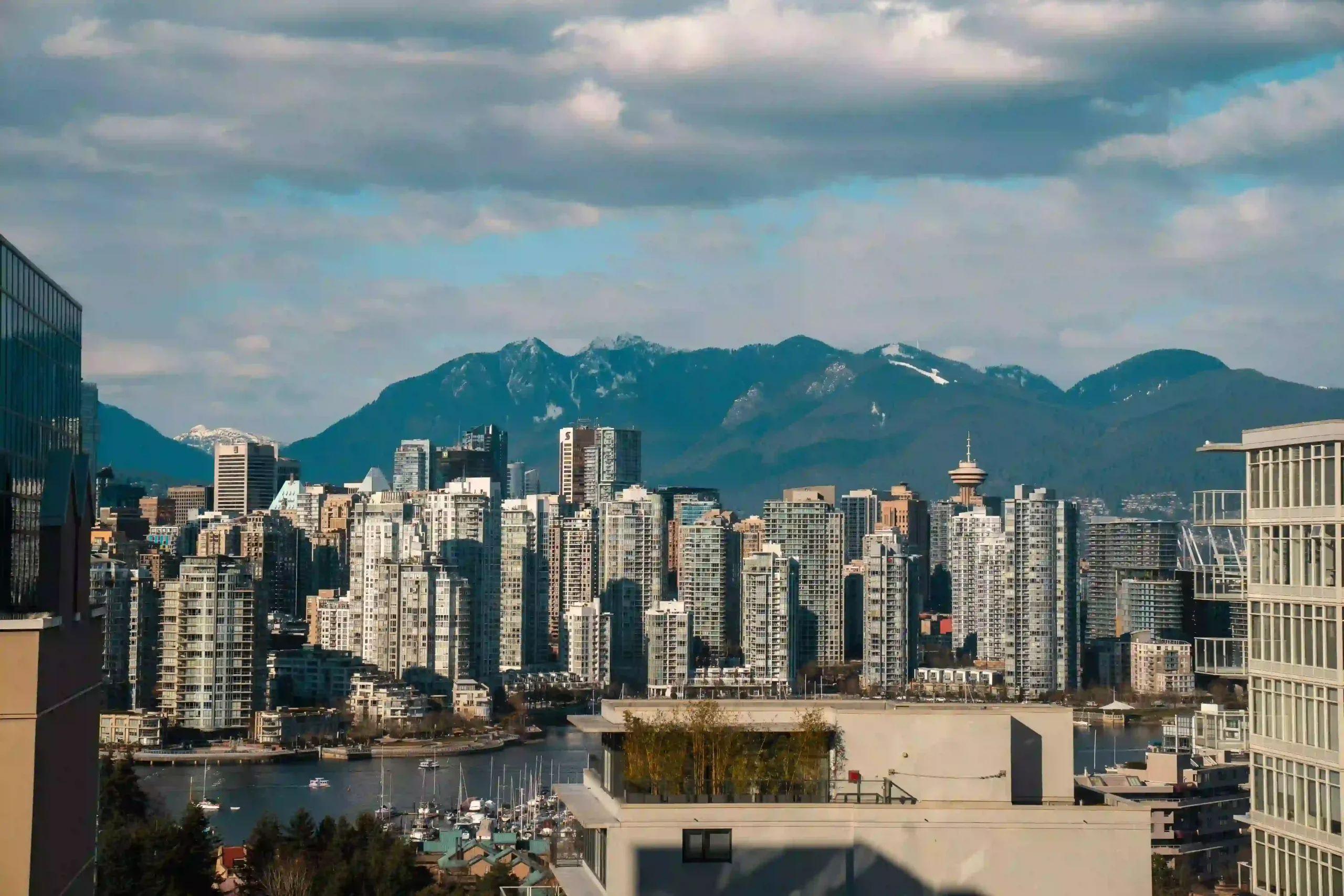 View of the Vancouver skyline featuring glass skyscrapers and a Harbour, with a mountain backdrop and a cloudy sky.