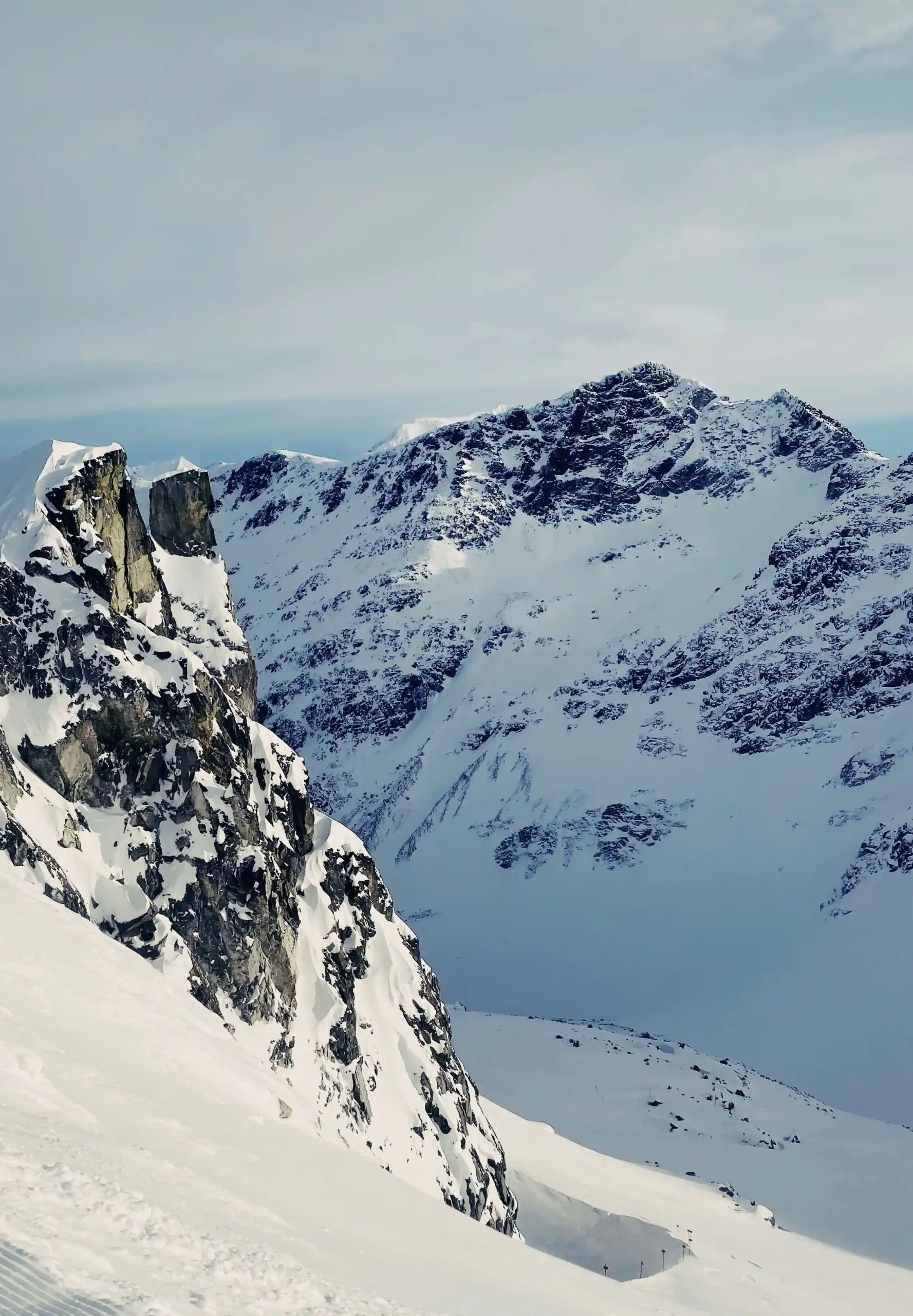 A vertical shot of a steep, snow-covered mountain face in Whistler Blackcomb. Dark, jagged rock formations jut out from the white snow in the foreground, with a massive, craggy peak rising behind it under a pale blue sky.
