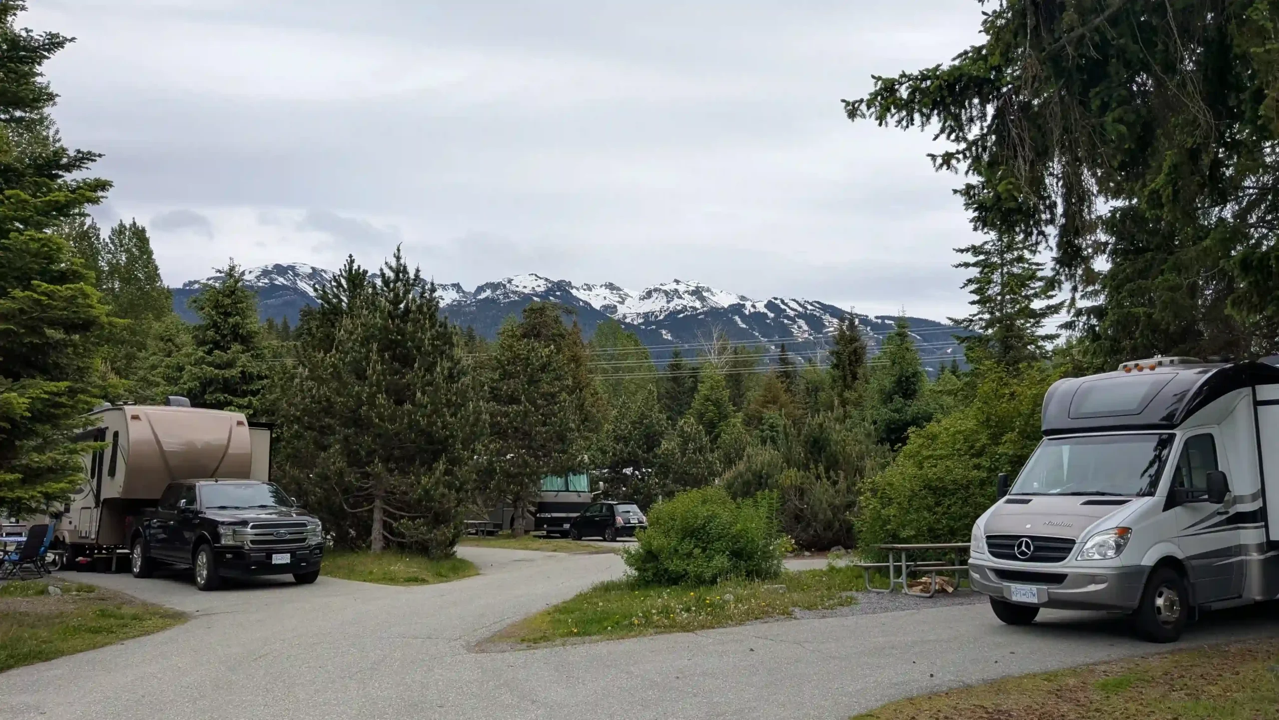 A paved campground area with several motorhomes tucked away. The site is surrounded by lush green pine trees, with the snow-capped peaks of the Whistler mountain range visible in the distance under an overcast sky.