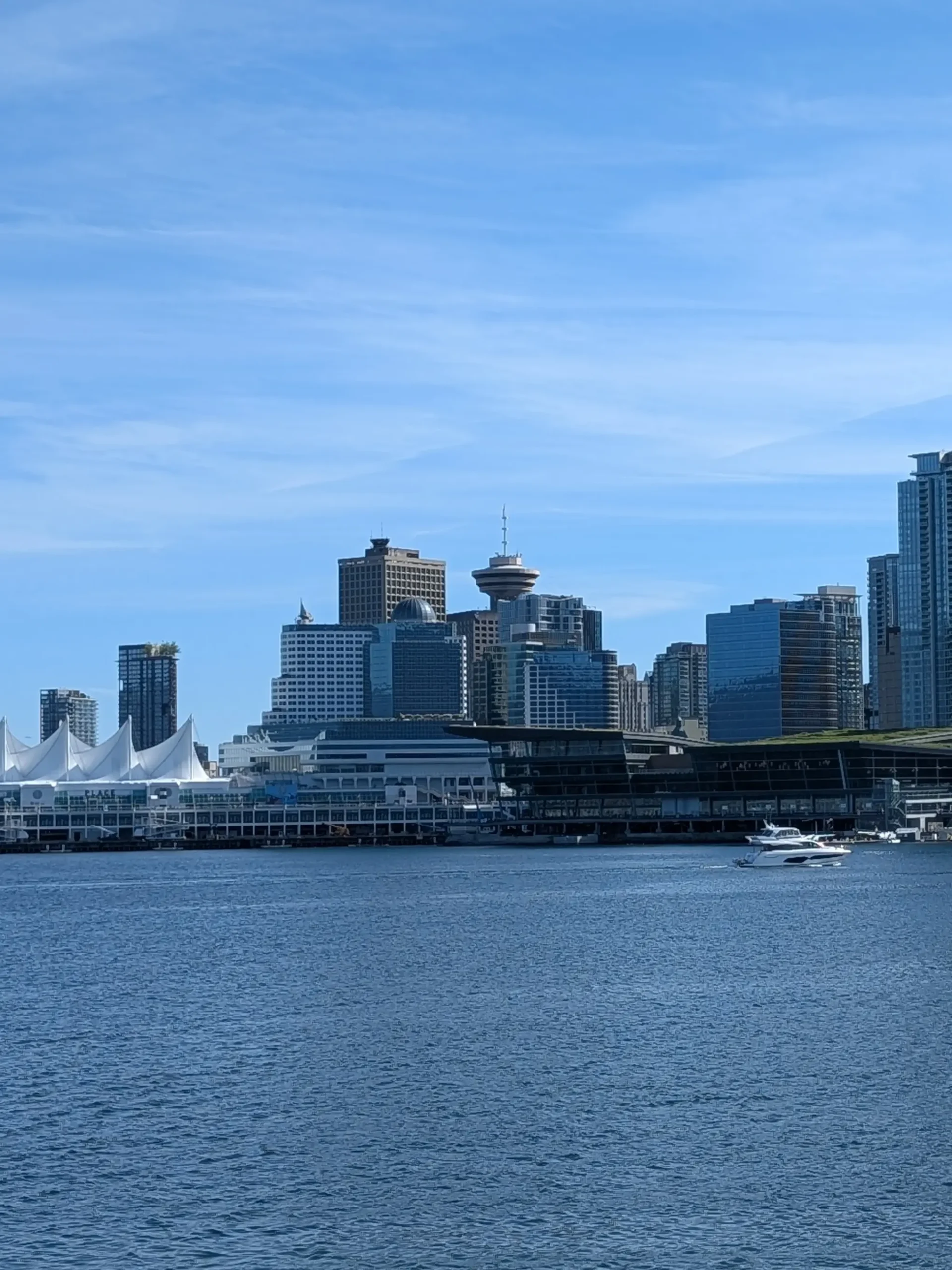 A vertical view across Burrard Inlet towards the Vancouver skyline, featuring Canada Place\'s white sails, the Vancouver Lookout tower, and modern skyscrapers on a clear sunny day.
