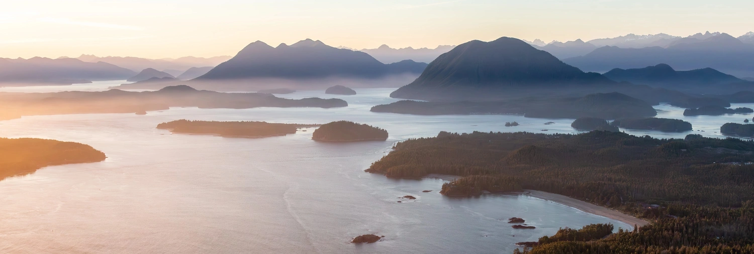Panoramic aerial view at golden hour showing forested islands, calm inlets, and sandy coastal beaches around Tofino.