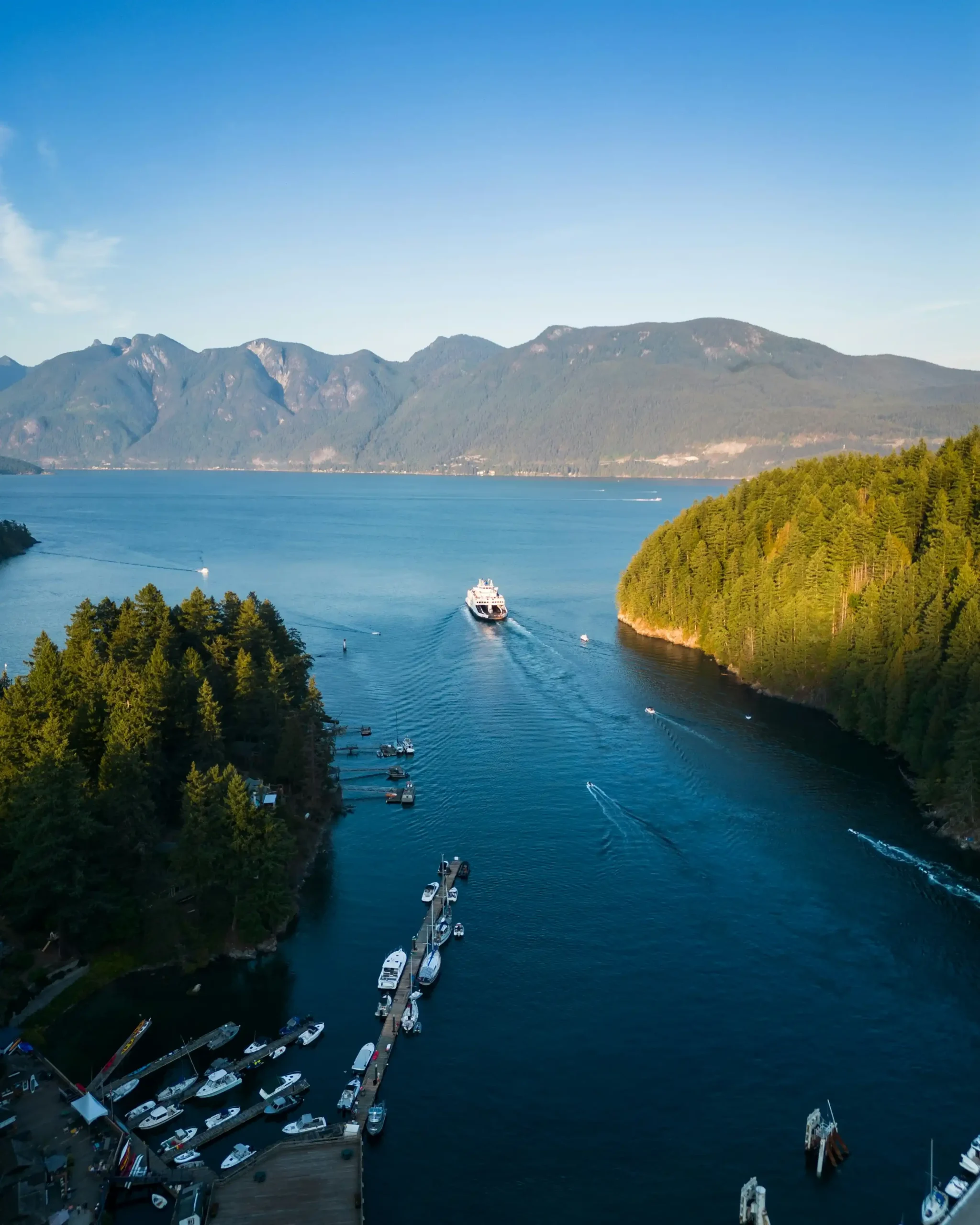 An aerial perspective looking down at a BC Ferry navigating deep blue waters between forested peninsulas departing Horseshoe Bay.