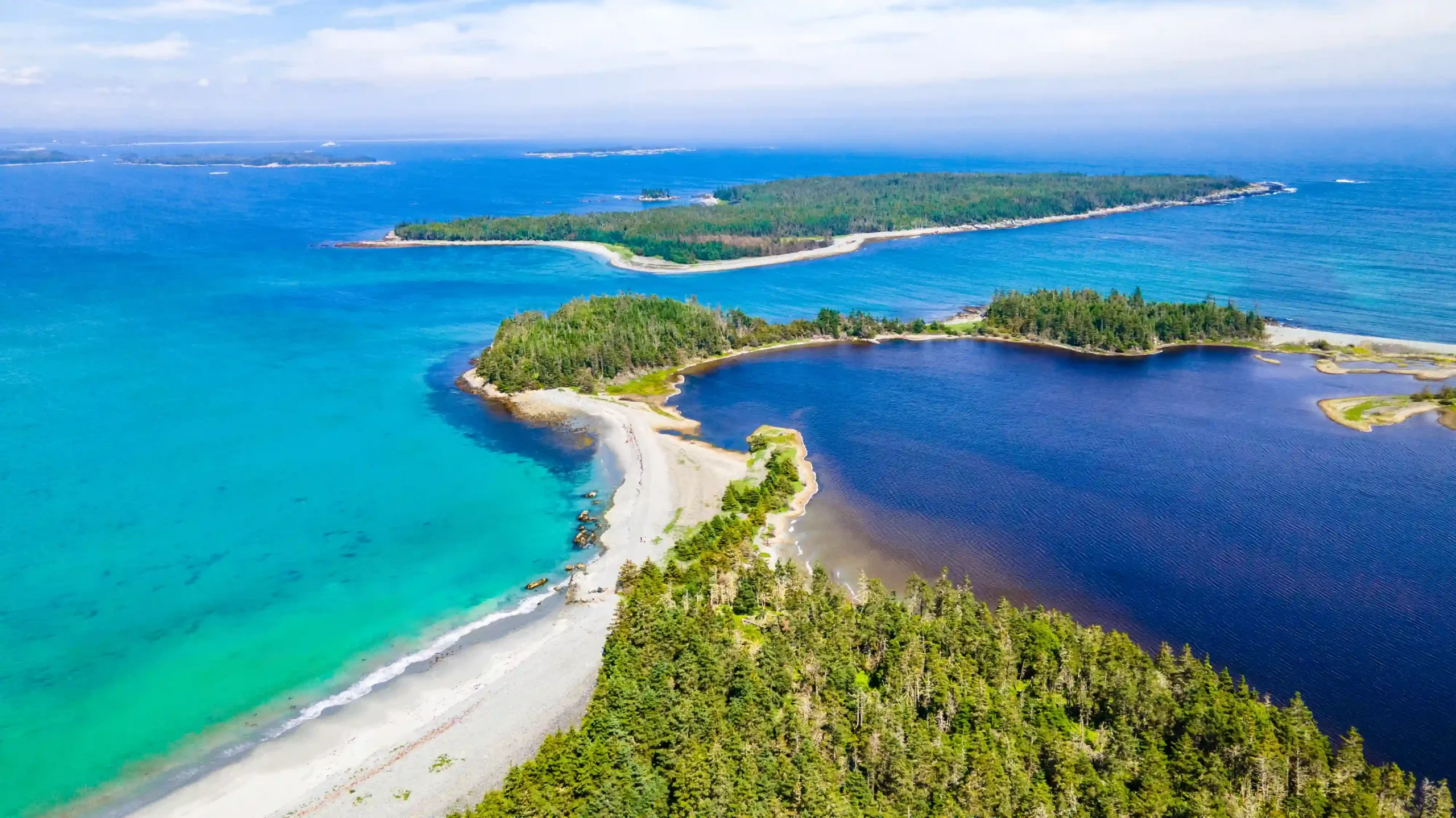 An aerial shot of Taylor Head Provincial Park in Nova Scotia, showing a narrow white sand beach separating bright turquoise ocean water on the left from a deep navy blue inland lagoon on the right. Lush green evergreen forests cover the rugged, rocky coastline under a clear blue sky.