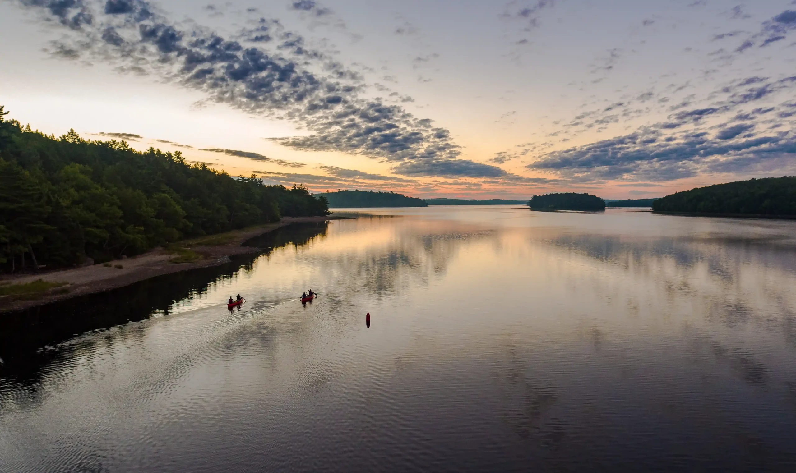 An aerial wide shot of Kejimkujik Lake at sunset. Two red canoes paddle across the calm, glassy water, leaving small wakes behind them. The sky is filled with soft purple and orange hues reflected in the water, framed by dense green forests on the shoreline under a patterned layer of clouds.