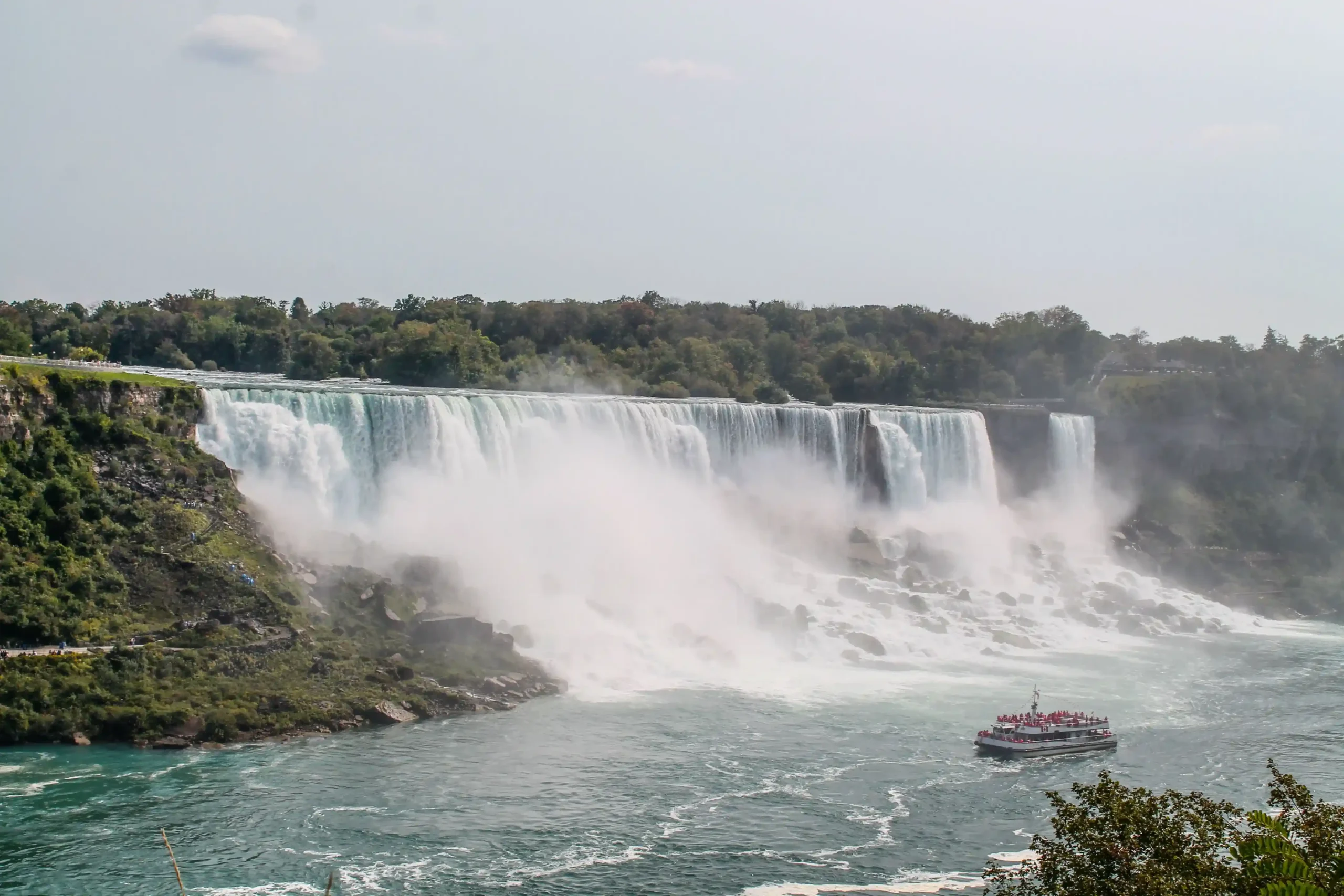 A tour boat filled with passengers in red ponchos sits in the turquoise water at the base of the falls, surrounded by white mist and spray.