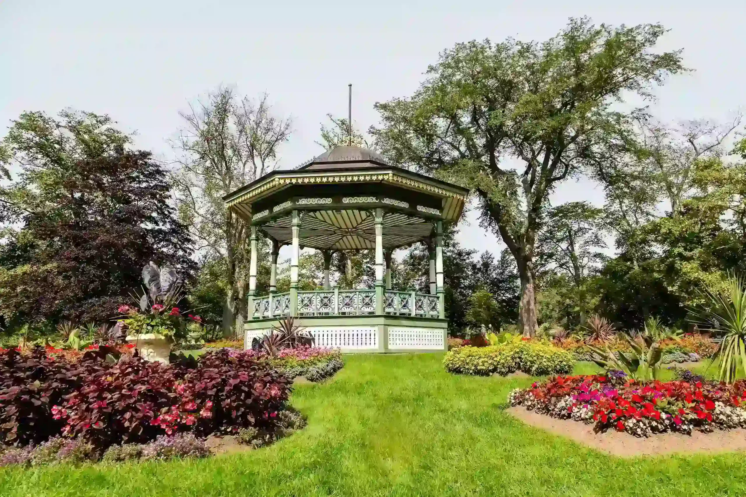 A bandstand is surrounded by vibrant flower beds containing red, purple, and yellow blooms, green grass, and large leafy trees under a bright sky.