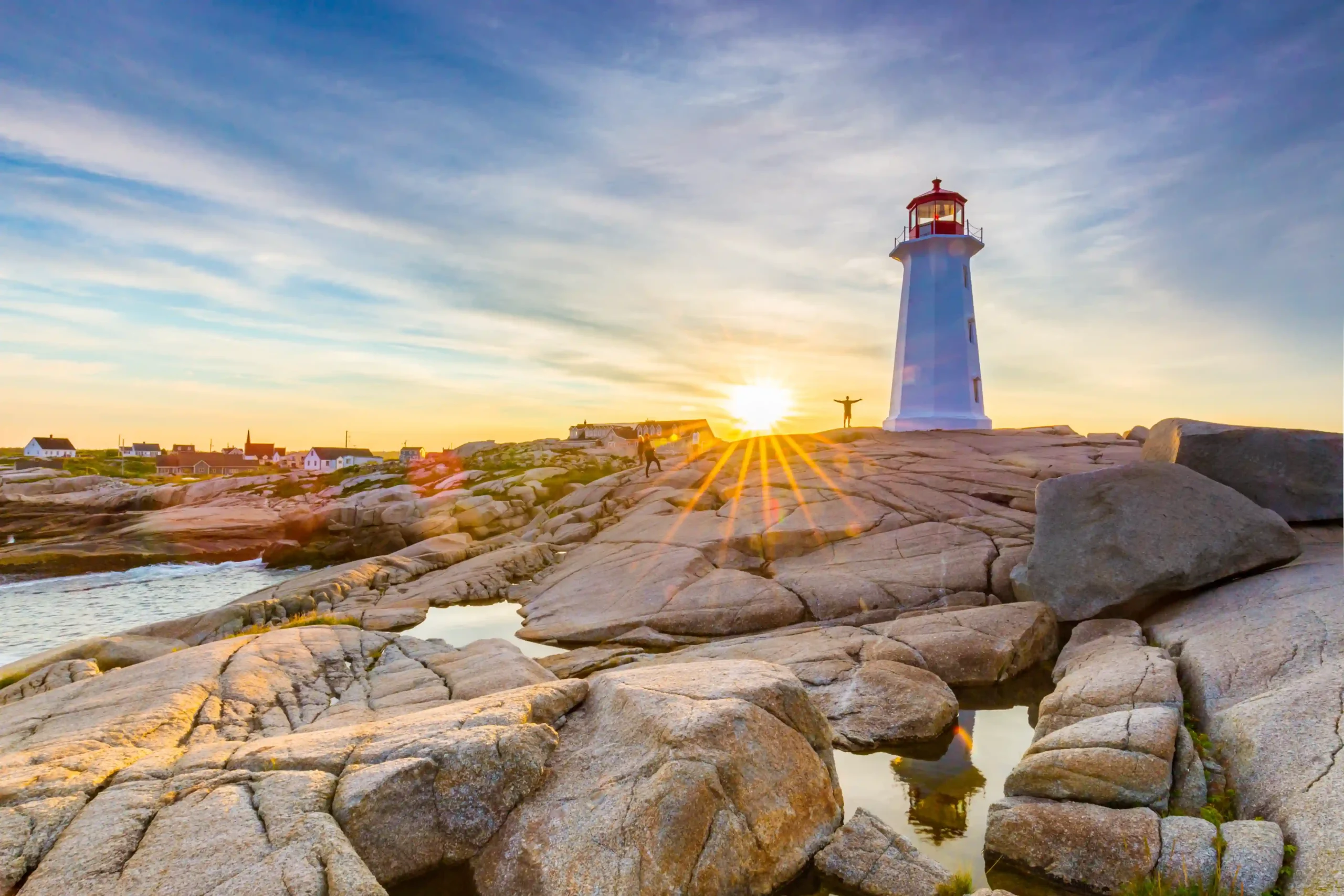 The sun is low on the horizon, creating a bright starburst effect and casting a warm golden glow over the massive, rounded granite rocks in the foreground. The iconic white lighthouse with its red lantern room stands tall against a sky streaked with soft blue and orange clouds.