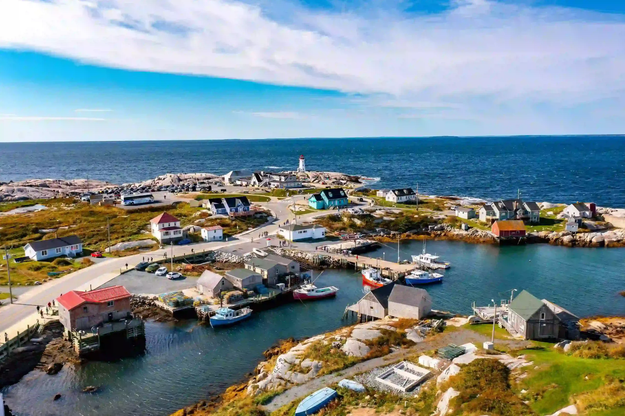 In the foreground, colorful fishing boats sit in a protected blue inlet surrounded by rustic wooden shacks. Beyond the village, the famous white and red Peggy\'s Point Lighthouse stands on massive, smooth gray granite rocks overlooking the deep blue Atlantic Ocean.