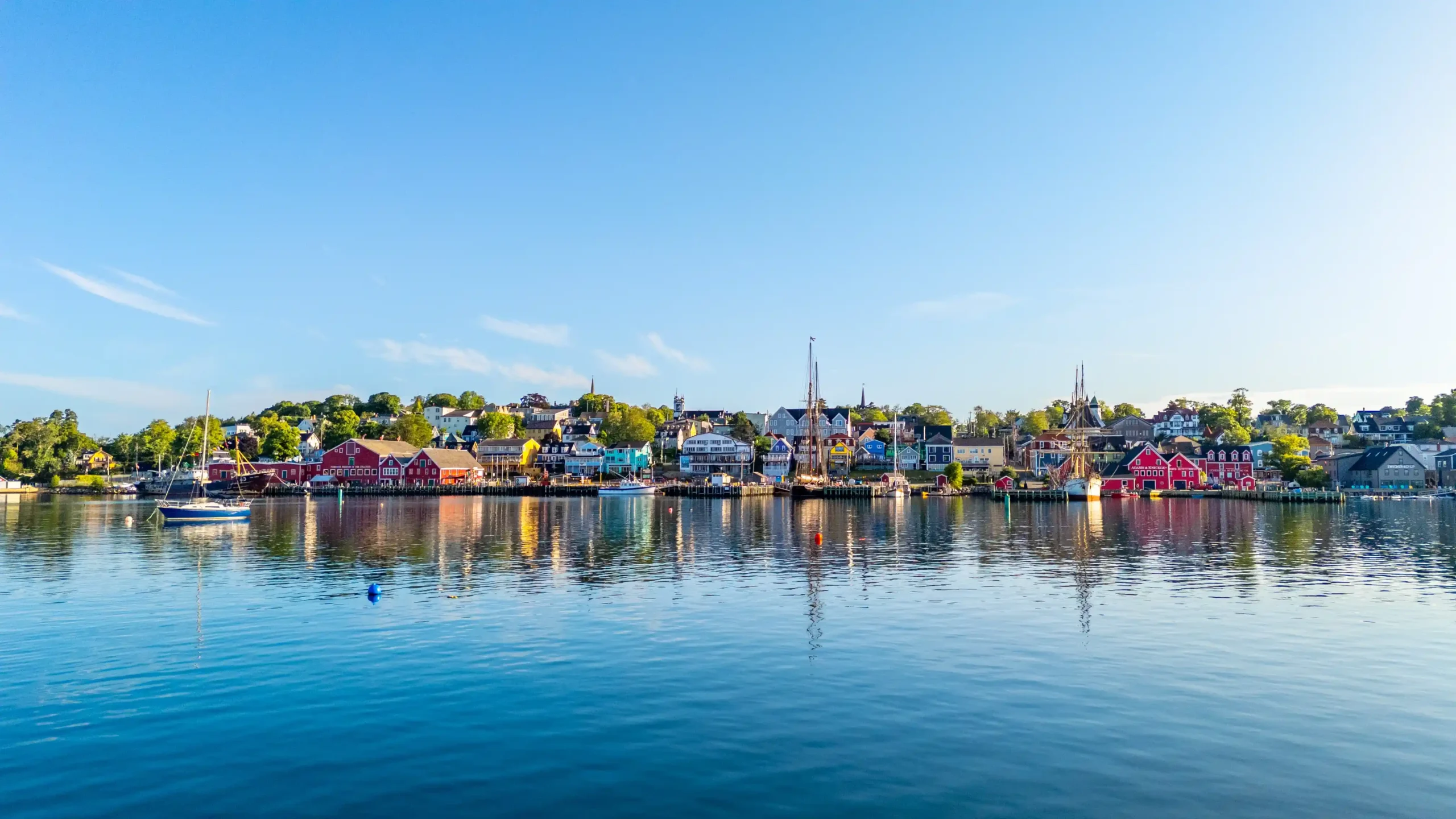 A wide-angle landscape photograph of the historic waterfront of Lunenburg, Nova Scotia. The calm blue water of the harbor perfectly mirrors the brightly painted, colorful wooden buildings (reds, blues, yellows) lining the shore. Several boats, including large sailing schooners, are moored at the docks under a clear, sunny blue sky.