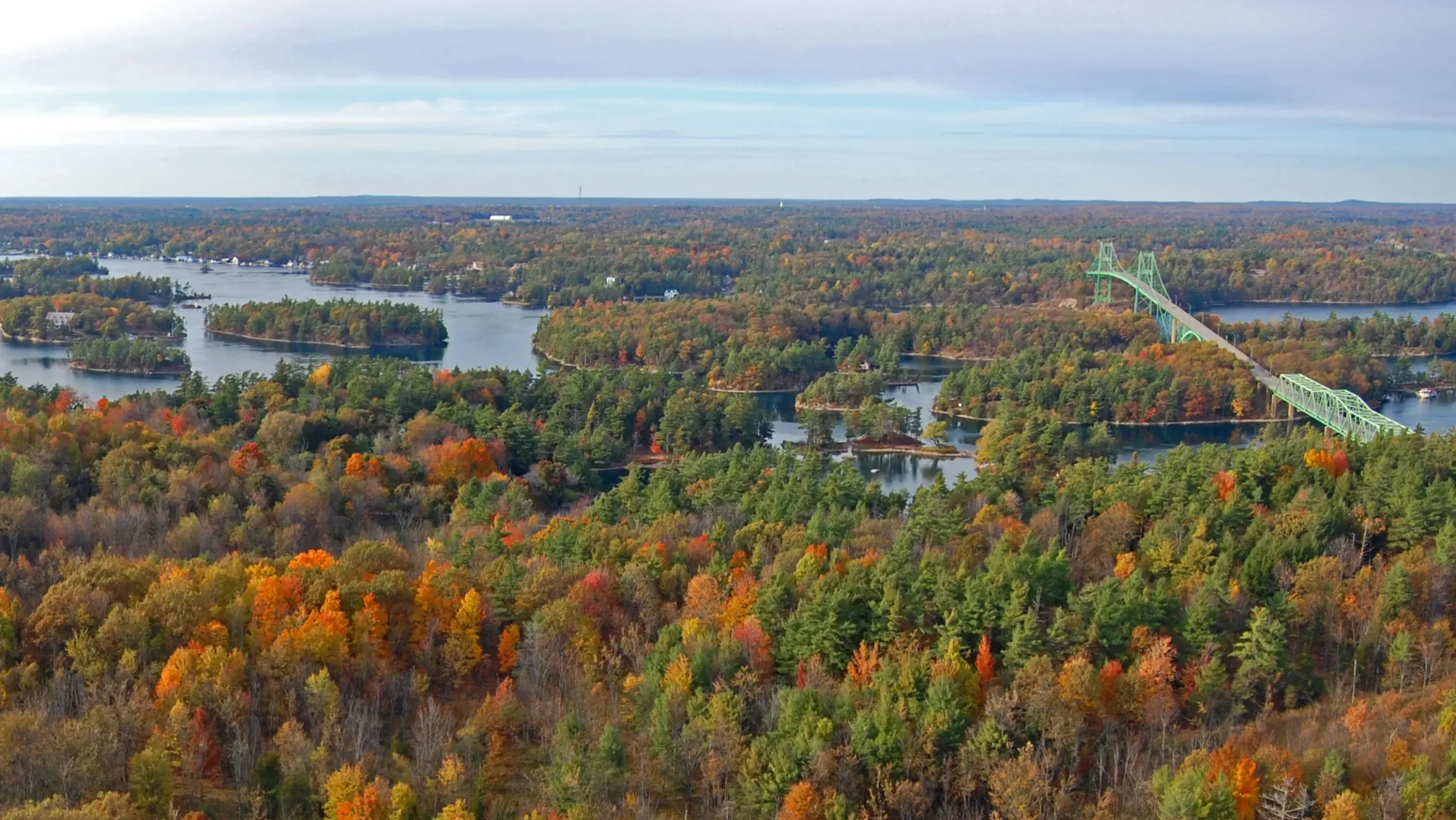 Aerial view of the Thousand Islands region on the St. Lawrence River during autumn, featuring a green suspension bridge connecting forested islands with vibrant red, orange, and yellow foliage.