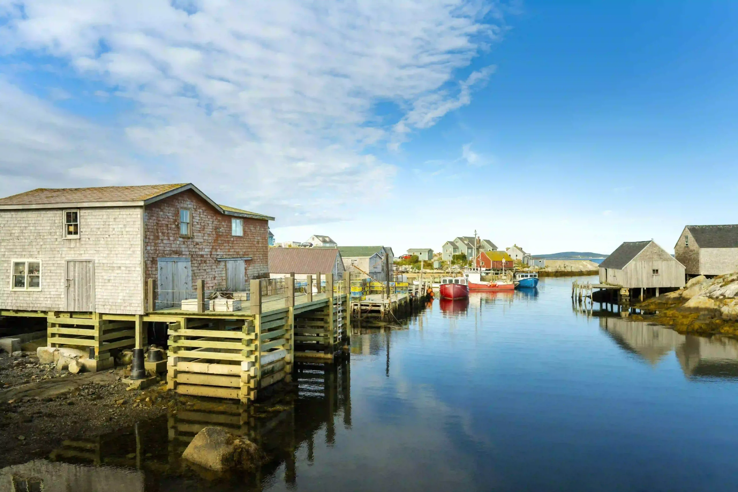 Weathered wooden fishing shacks on the water\'s edge, reflecting in the still blue surface. Small, colorful fishing boats are docked in the background under a bright blue sky.