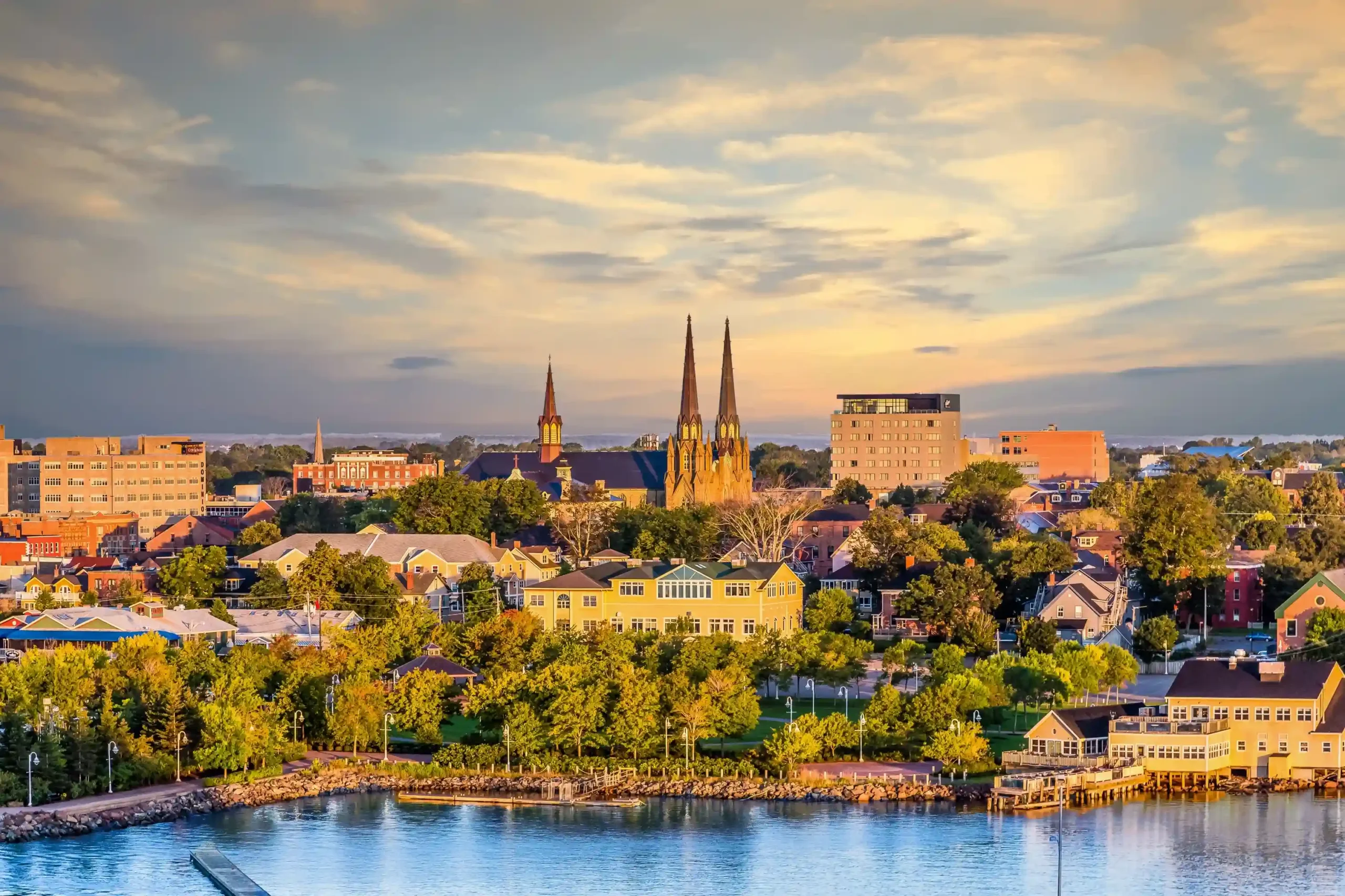 View of the Charlottetown, Prince Edward Island skyline at sunset, featuring the twin spires of St. Dunstan\'s Basilica, colorful buildings, lush green trees, and the calm blue water of the harbour.