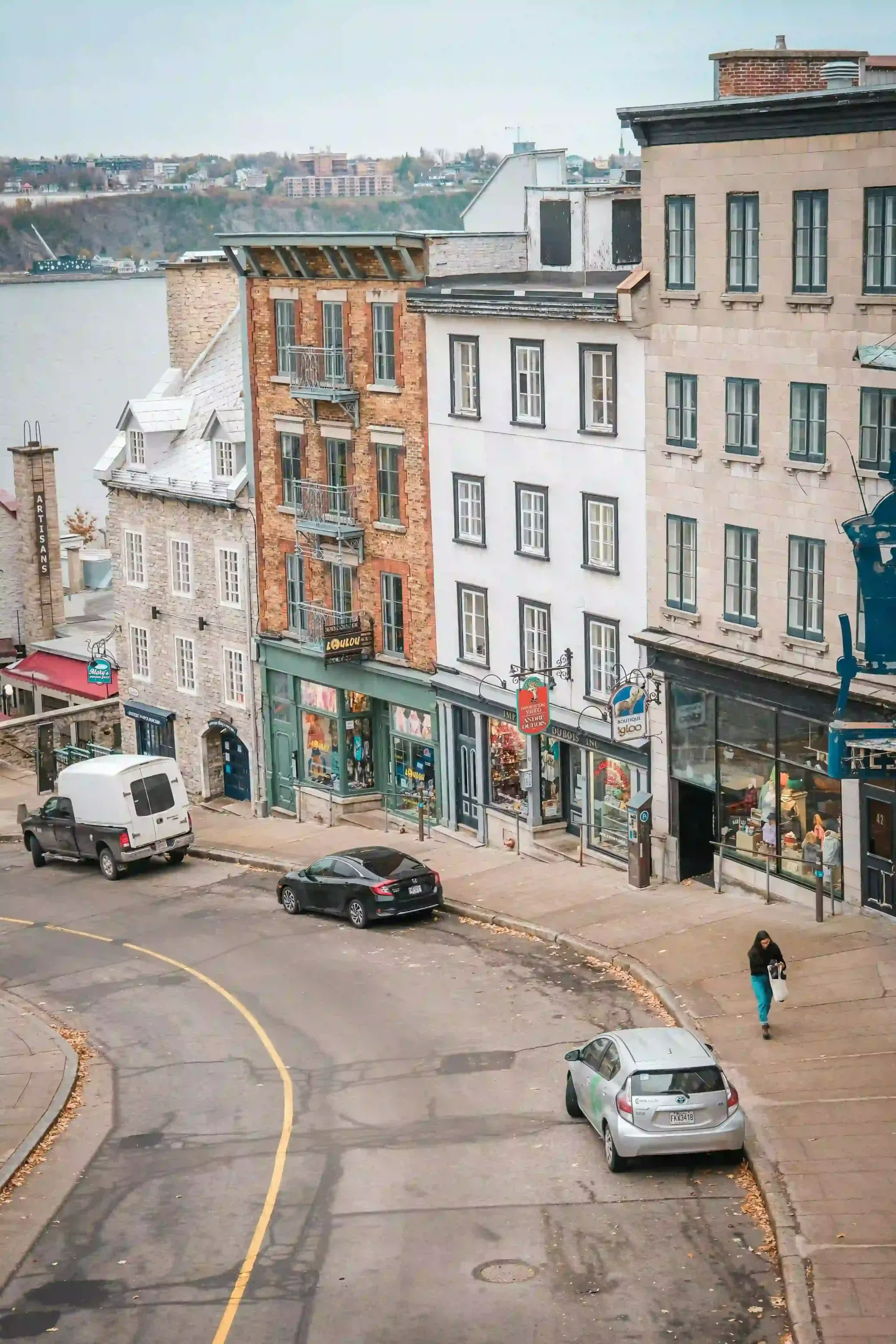 A high-angle view of a curved street in Quebec featuring stone and brick buildings with storefronts, overlooking the St. Lawrence River in the background.