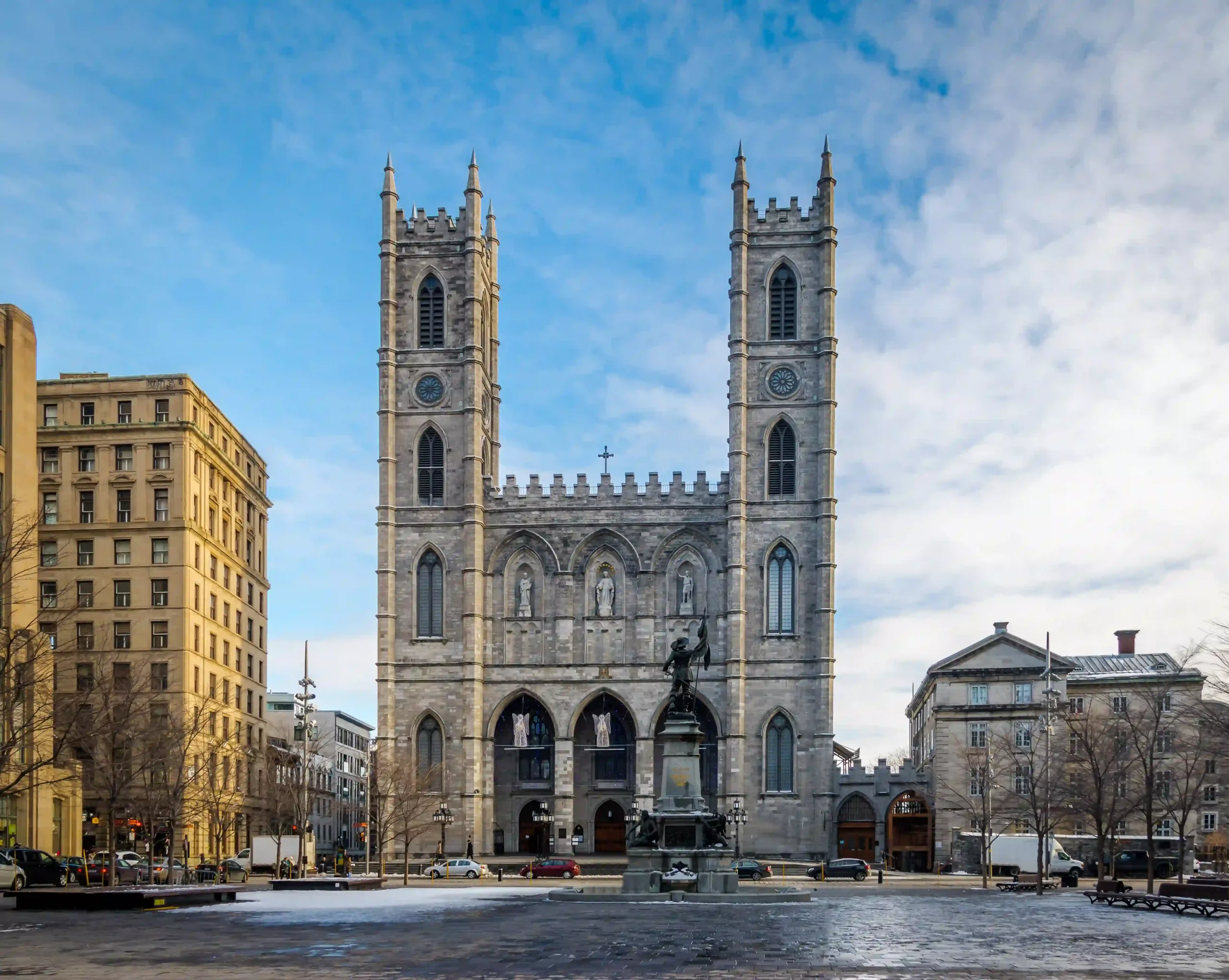 View of the Notre-Dame Basilica in Montreal, Quebec, featuring two twin towers and the Maisonneuve Monument in the foreground at Place d\'Armes under a bright blue sky with light snow on the ground.