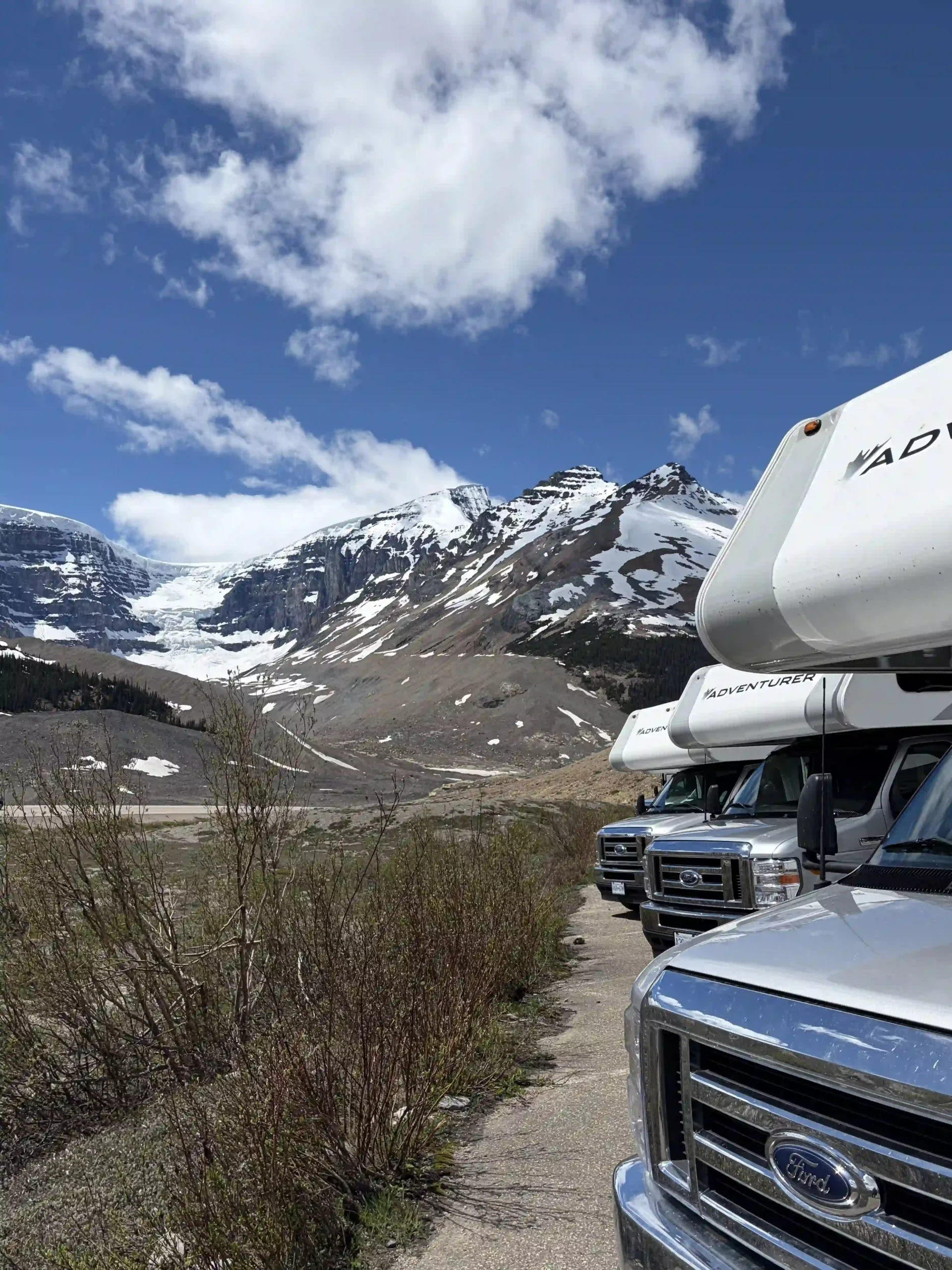 A row of white Adventurer motorhomes parked along a roadside overlook, with a stunning backdrop of snow-capped mountains and a flowing glacier under a blue sky.