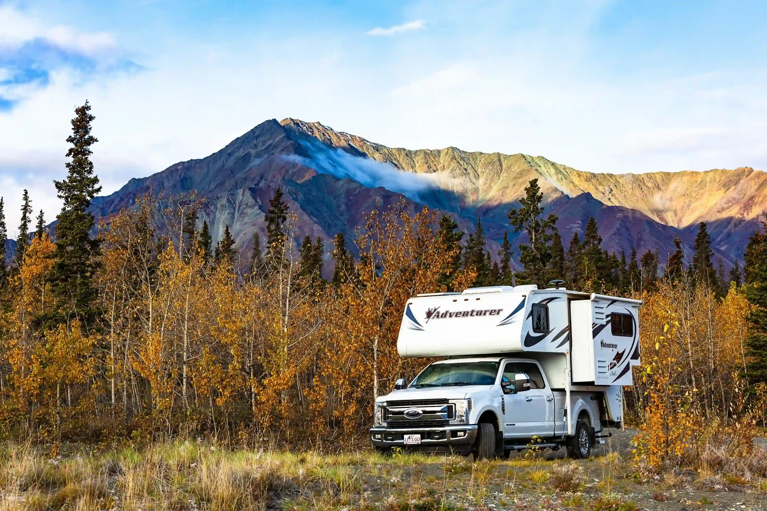 A white truck camper parked on a field surrounded by autumnal yellow-leafed trees.