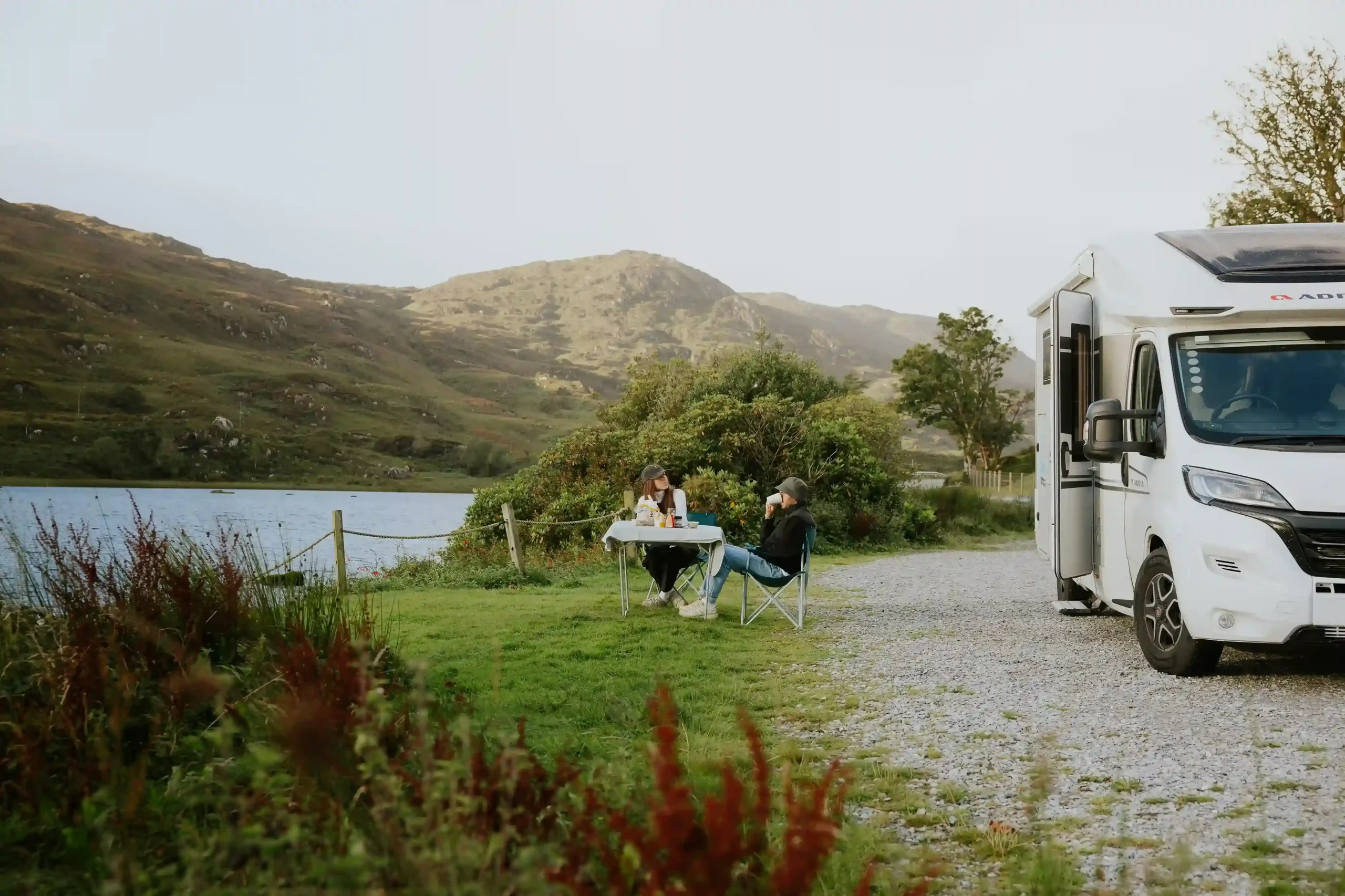 A couple sits at a folding table outside their motorhome, enjoying a meal by a lake surrounded by rolling green hills.