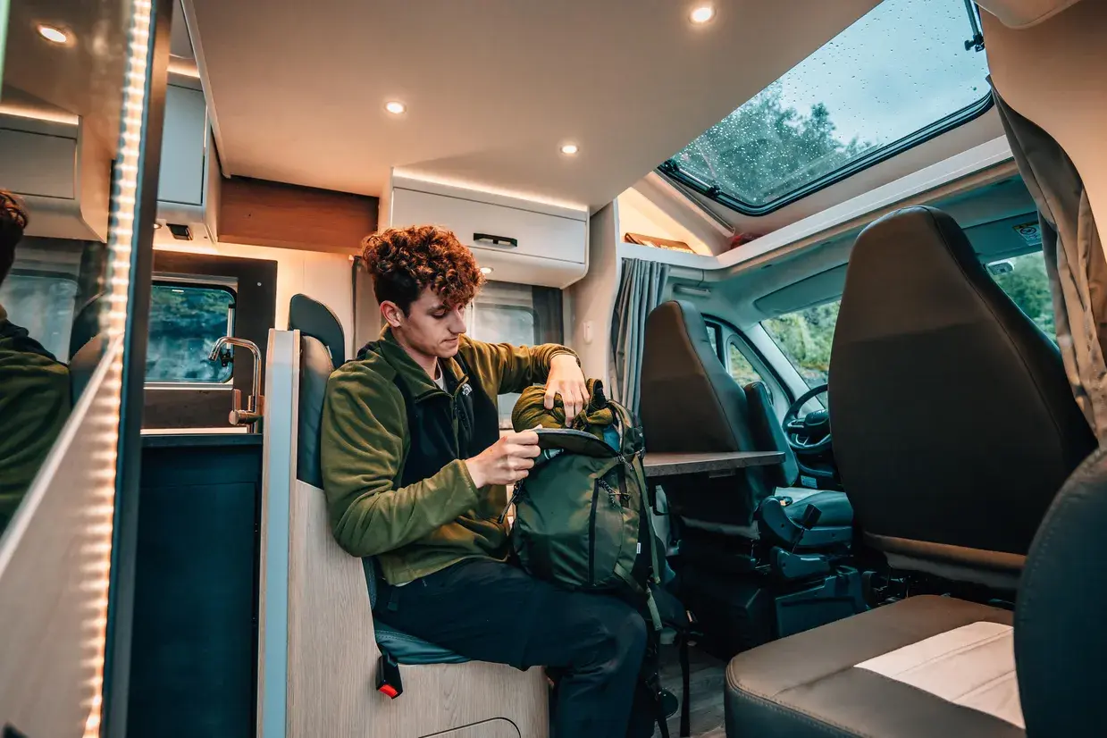 Interior view of a modern motorhome where a young man sits and packs a green hiking backpack.