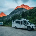 Two white camper vans are parked on a gravel lot nestled in a valley. The sun strikes the surrounding steep, rocky mountain peaks, illuminating them with a bright orange-red glow against a cool, blue-toned sky.