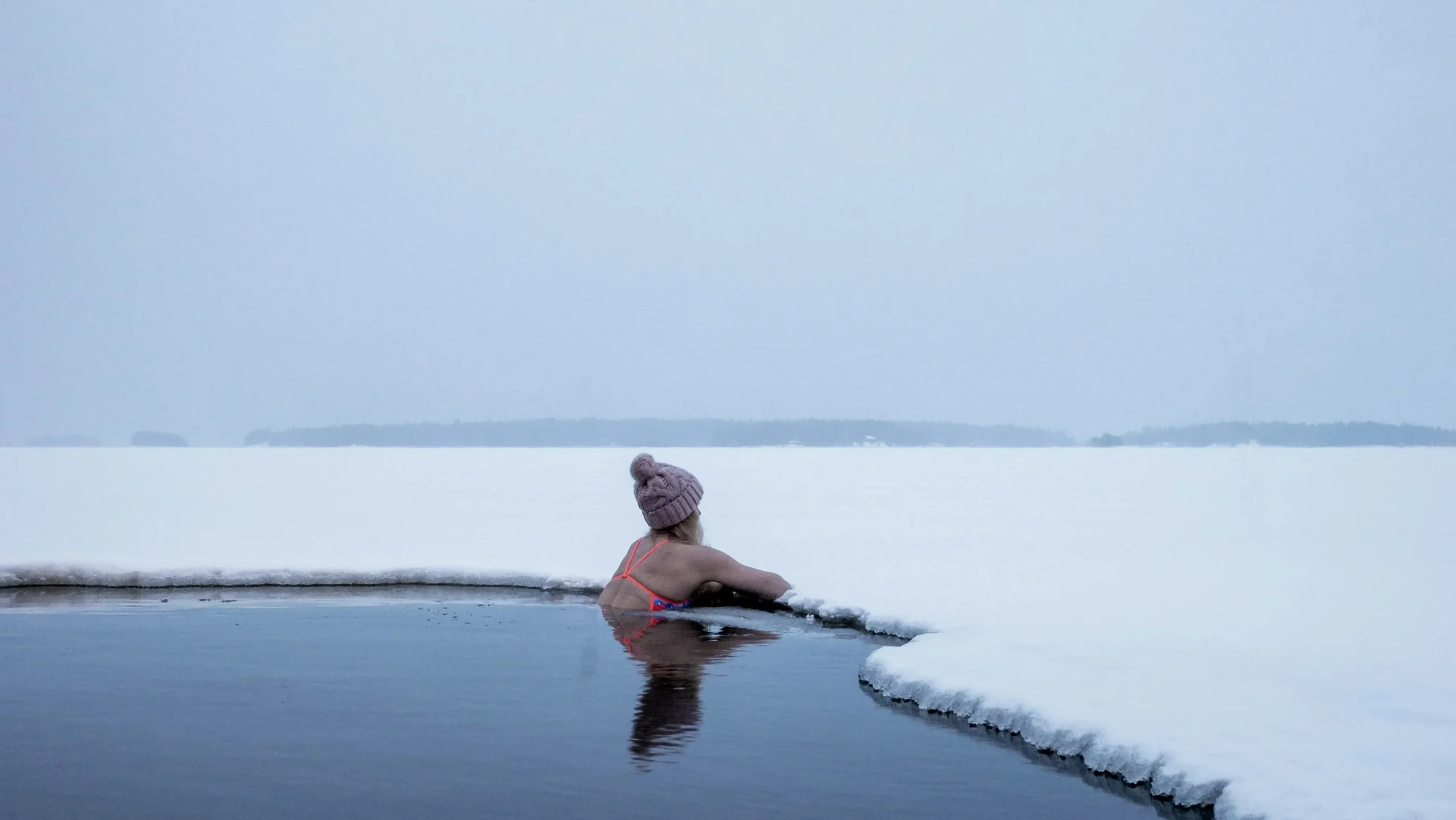 A woman wearing a pink knit beanie and swimwear performs avantointi, immersed in a hole cut through a thick frozen lake in Finland.