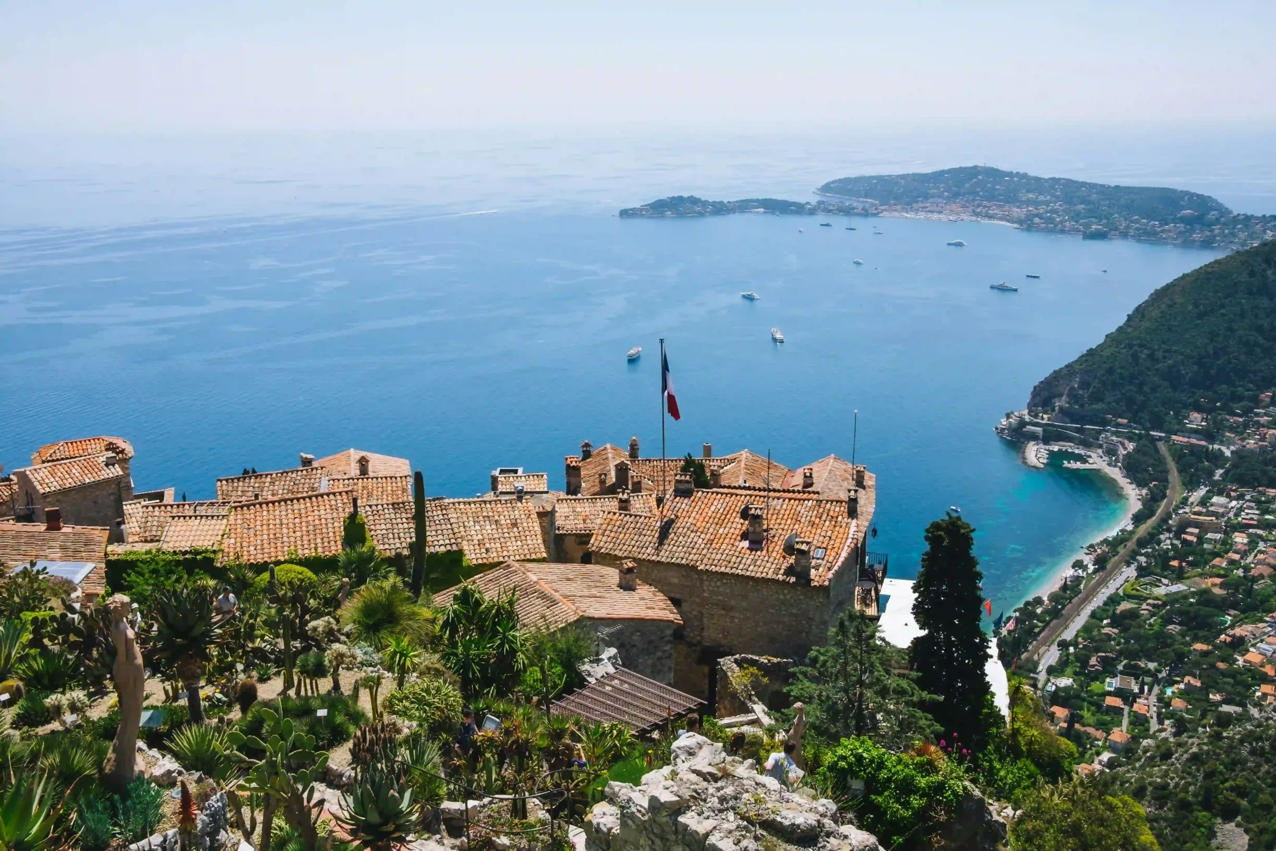 High-angle view of Èze, featuring terracotta rooftops, a French flag, and a sweeping vista of the deep blue Mediterranean Sea and coastline with boats.