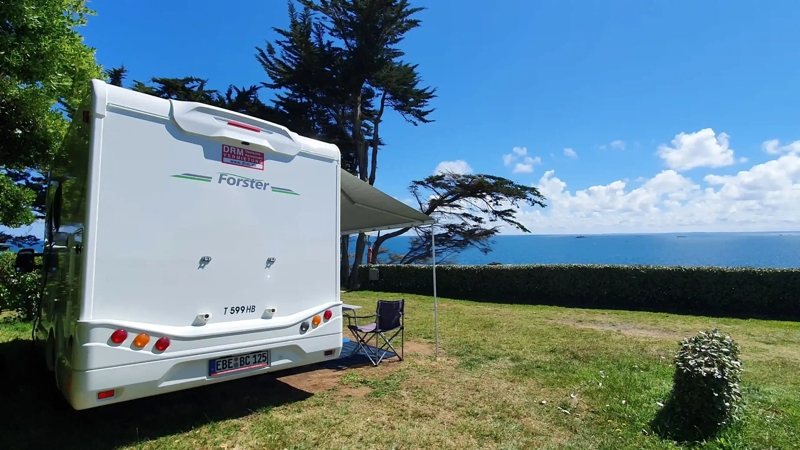 White motorhome parked on a grassy campsite pitch with its canopy extended and a camping chair set up, offering scenic views of the blue ocean.