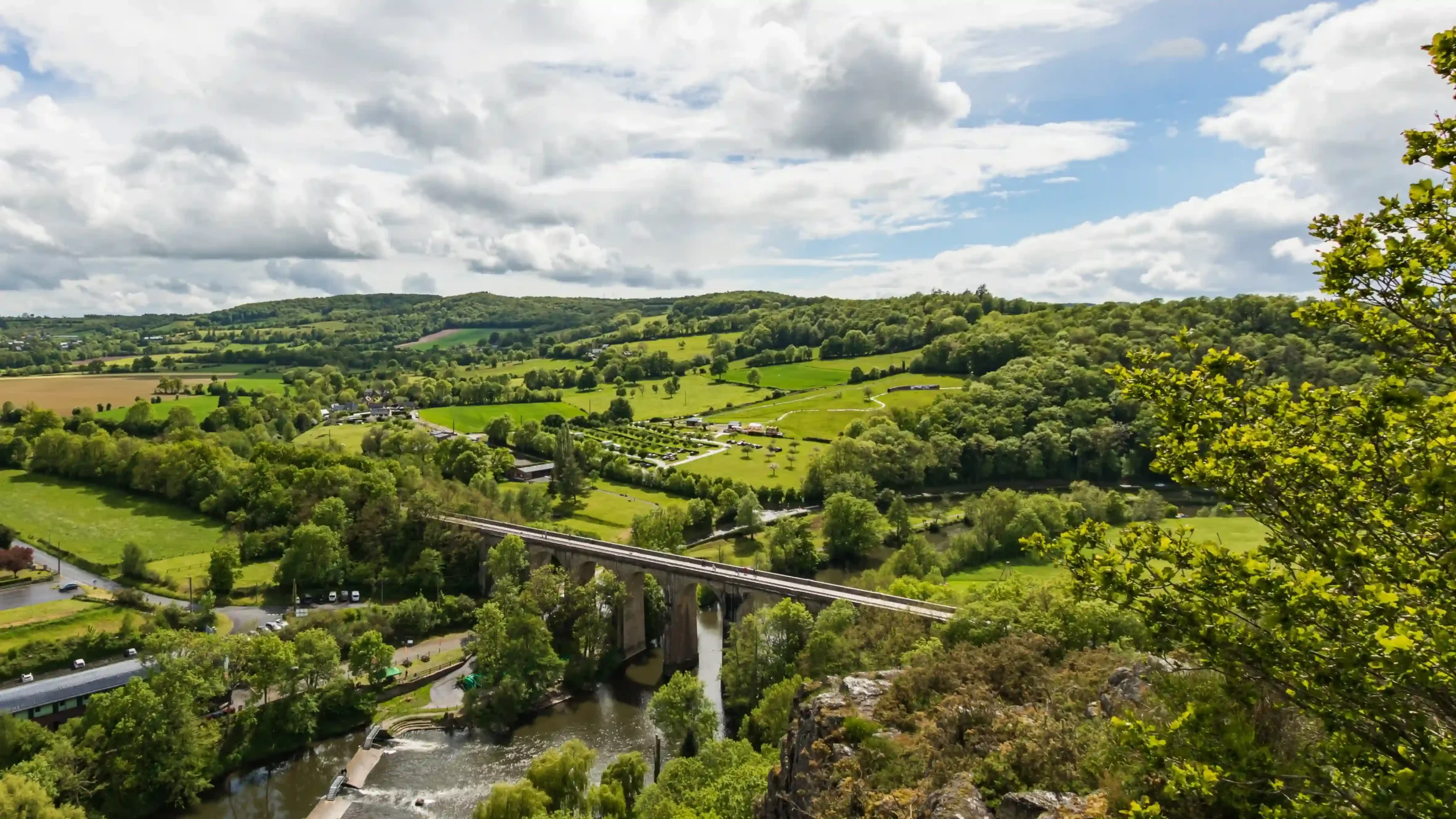 Elevated panoramic view of a stone railway viaduct crossing the Orne River amidst rolling green hills, dense foliage, and countryside pastures in Clécy.