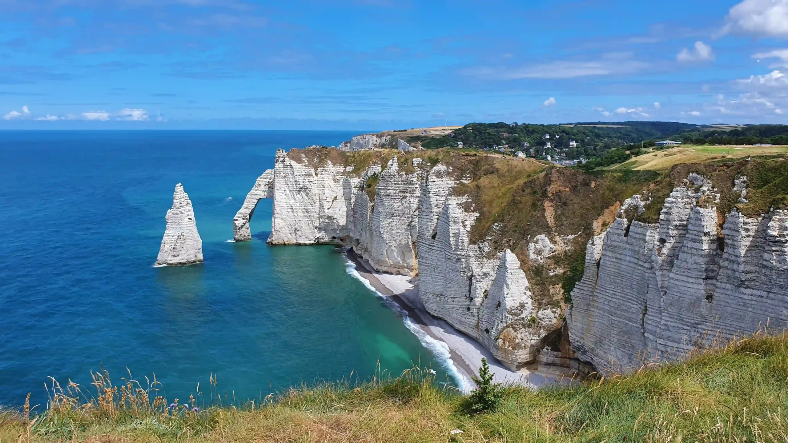 High-angle scenic view of the dramatic white chalk cliffs, natural rock arch, and pointed sea stack (The Needle) in Étretat, Normandy, set against deep blue and turquoise sea under a sunny blue sky.