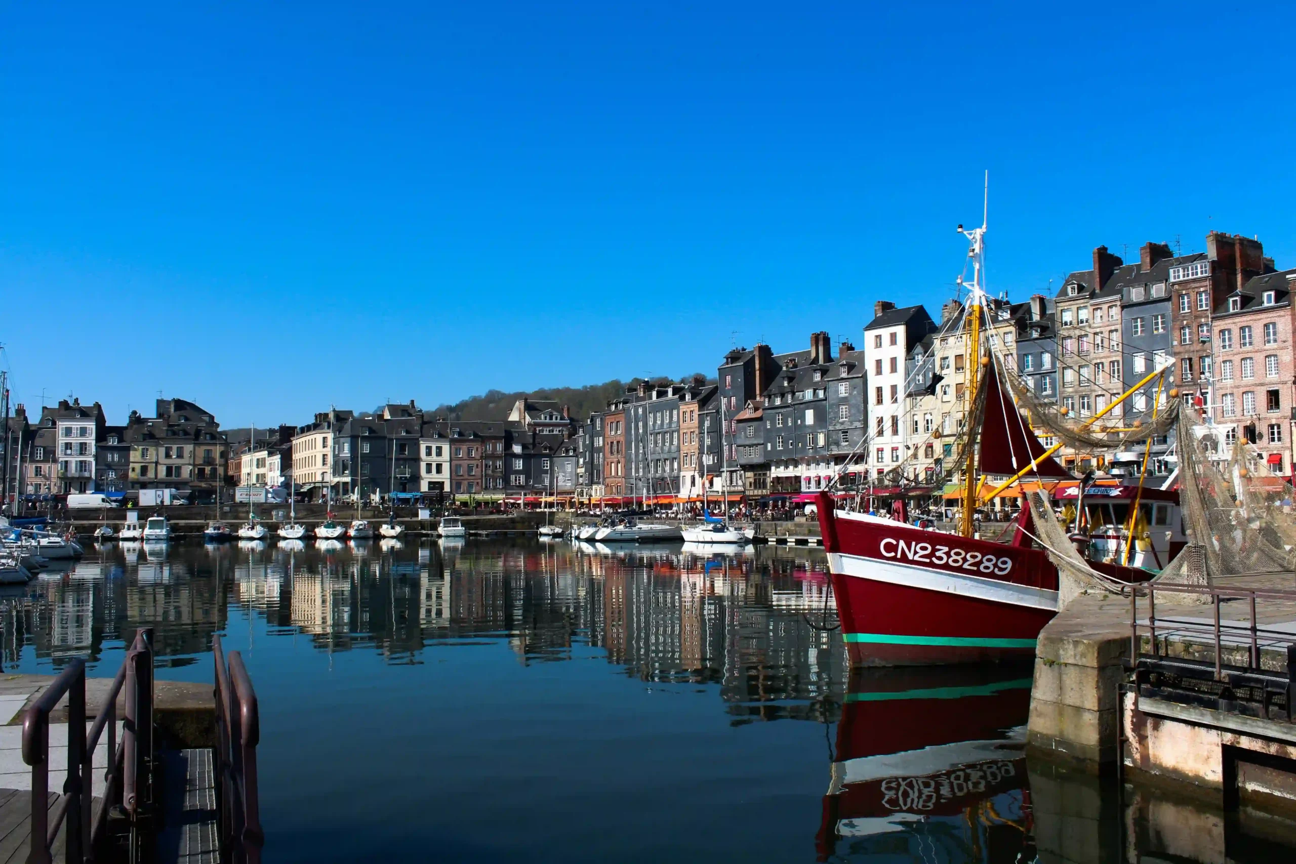 Scenic view of the calm water in Honfleur\'s historic harbour reflecting tall, slate-fronted buildings, with a red and white fishing trawler docked in the foreground under a vibrant clear blue sky.