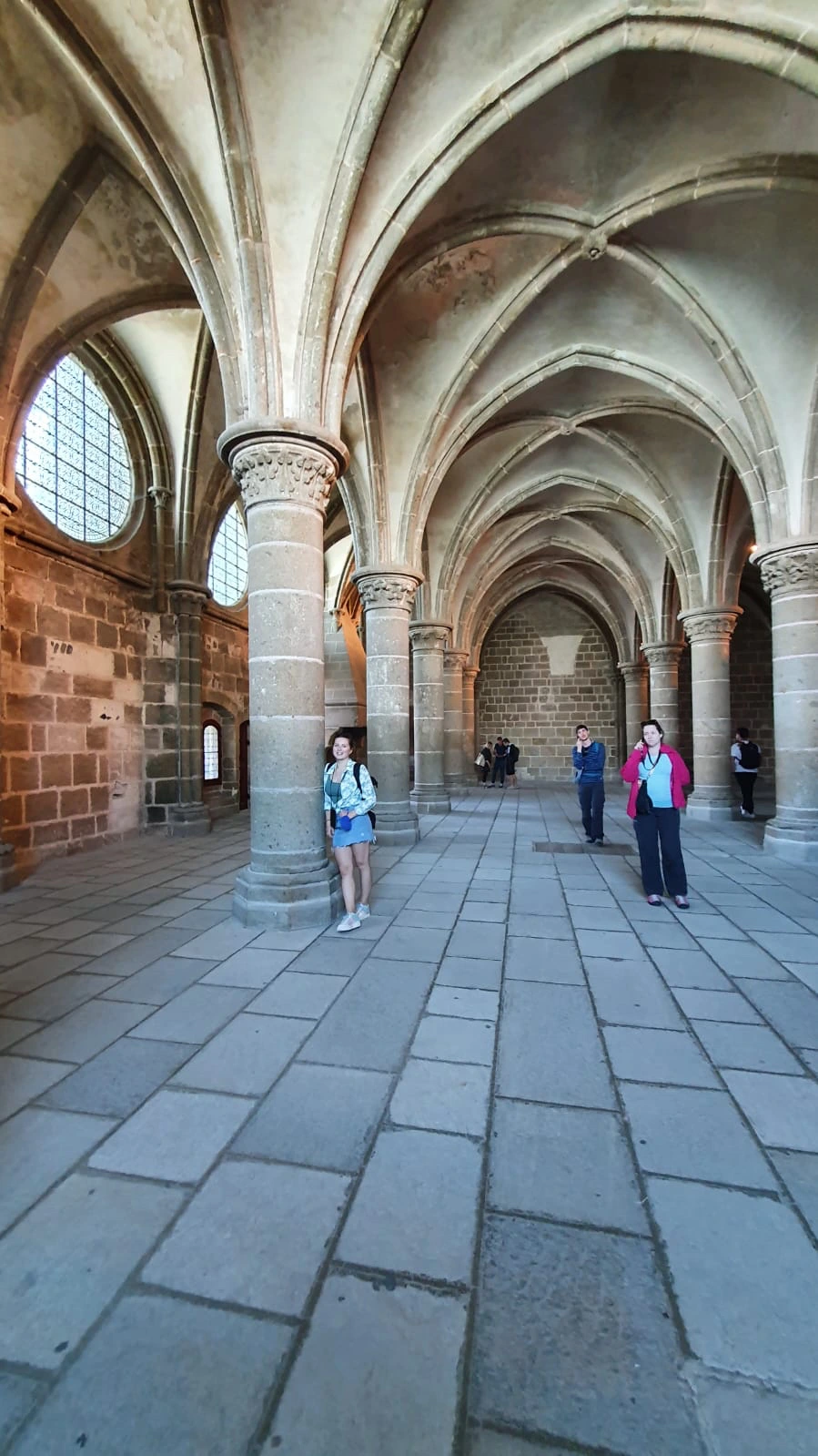 Spacious Gothic hall inside Mont-Saint-Michel Abbey featuring sturdy stone columns, ribbed vaulted ceilings, flagstone flooring, and visitors exploring the space.