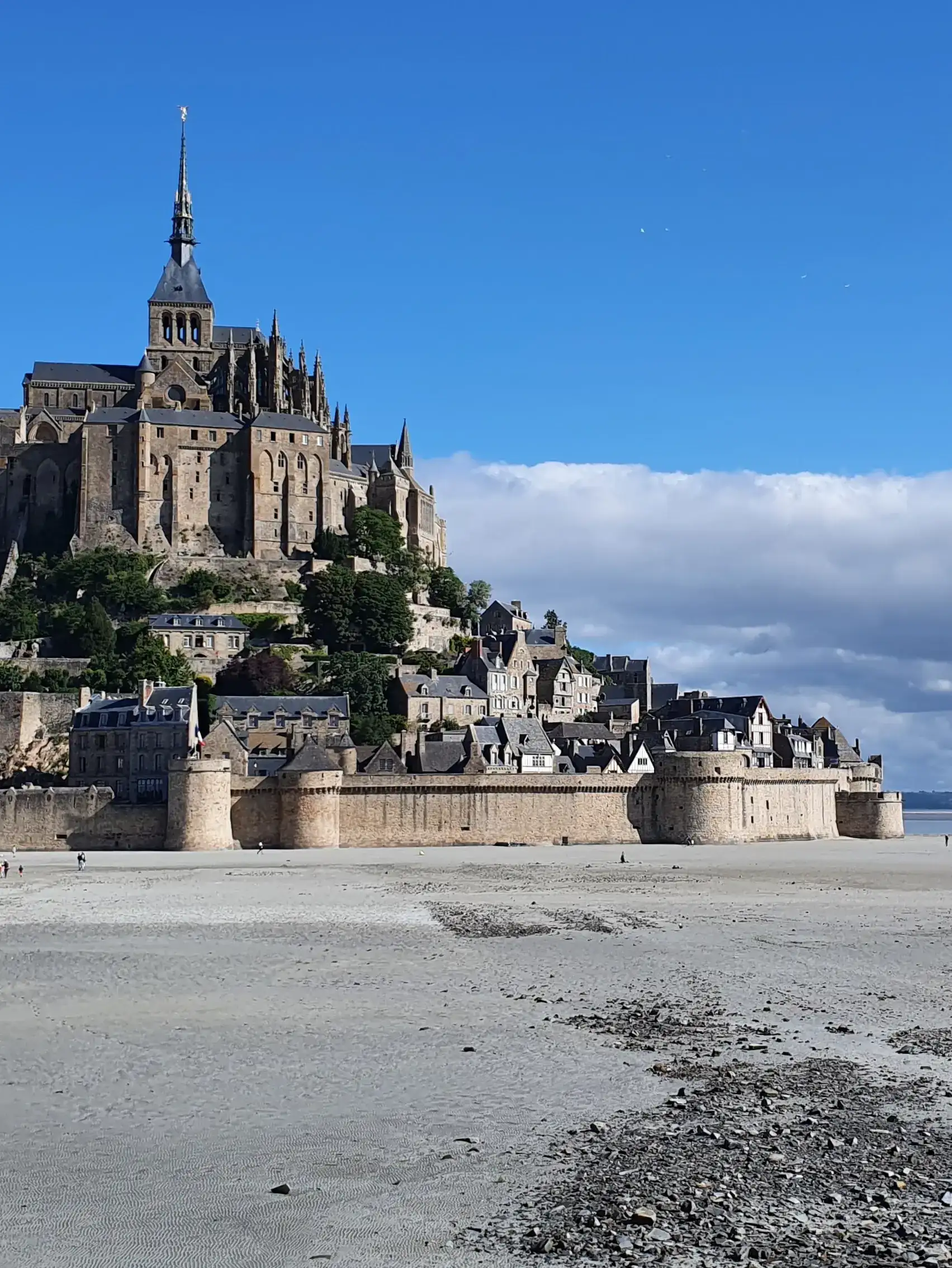 Low-angle view of the fortified stone walls of Mont-Saint-Michel standing above a wide sandy tidal flat under a clear blue sky.