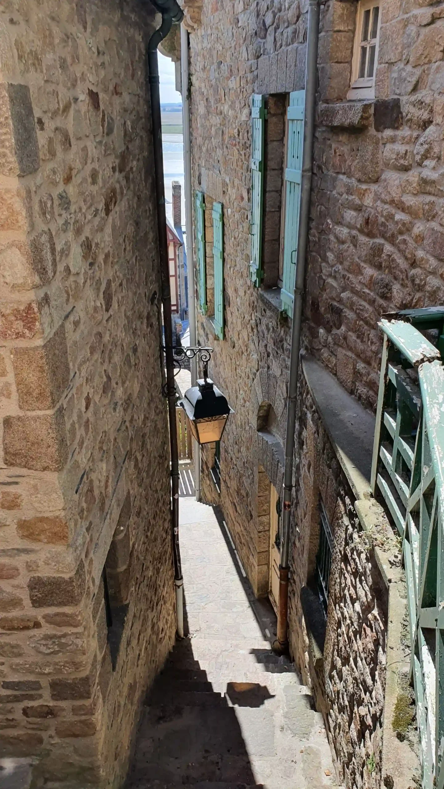 Looking down an extremely narrow medieval stone passageway with steps, a hanging lantern, and rustic stone walls.