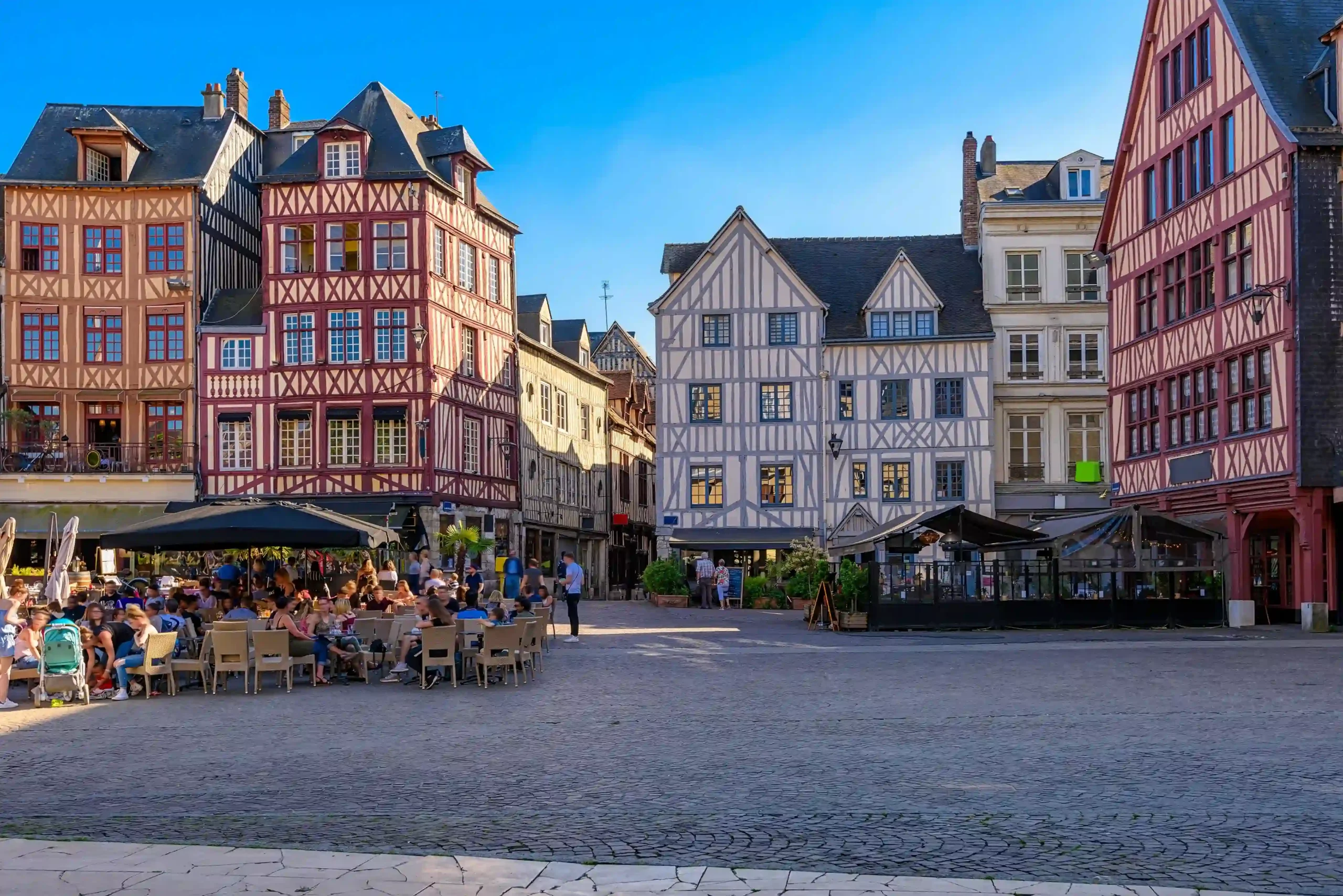 Sunlit historic town square surrounded by colorful medieval half-timbered buildings, featuring outdoor cafe dining terraces and people enjoying a bright day in Rouen.