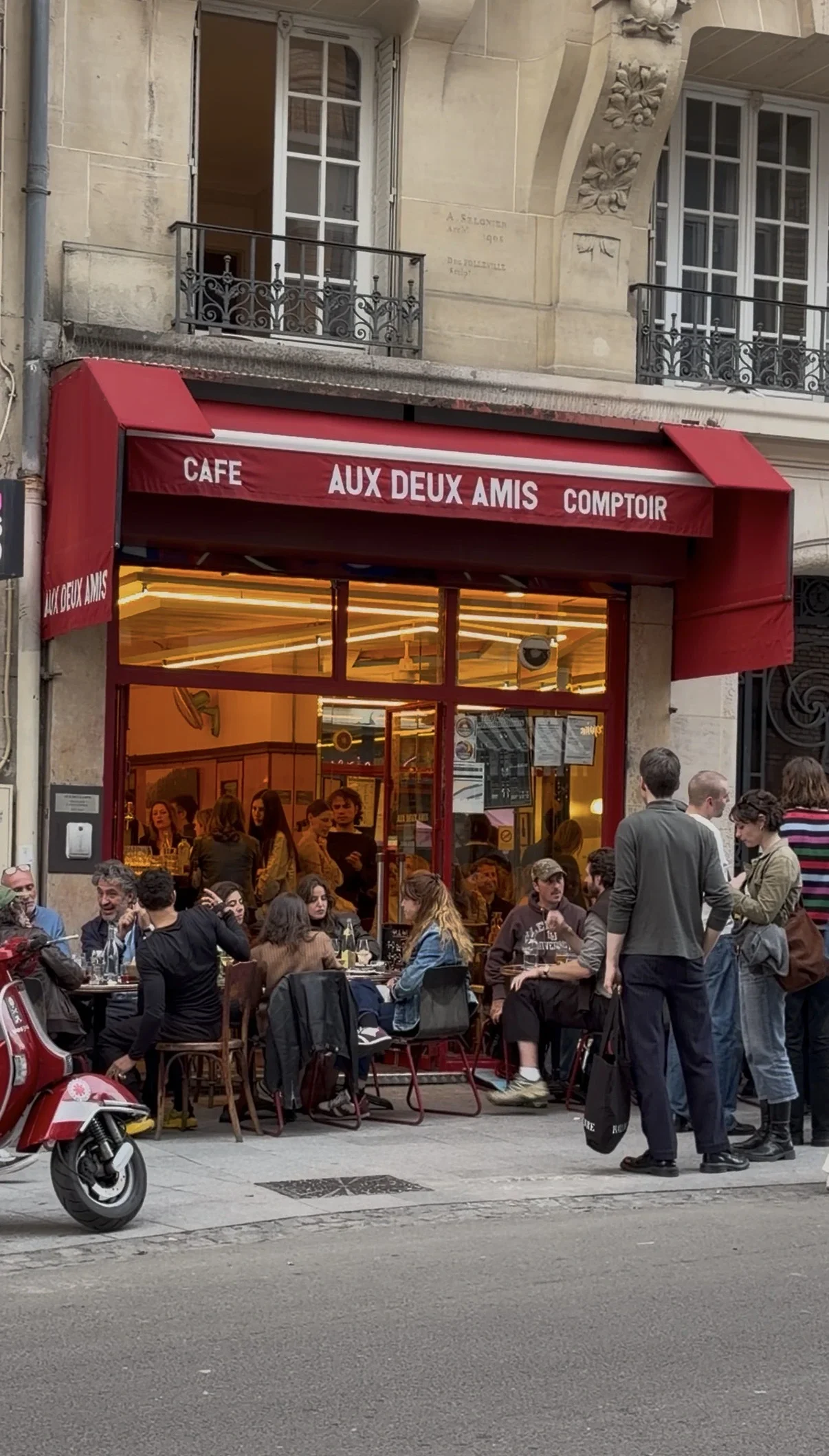 Lively outdoor terrace of a Café in Paris, showing people seated at tables under a bright red canopy alongside a street.