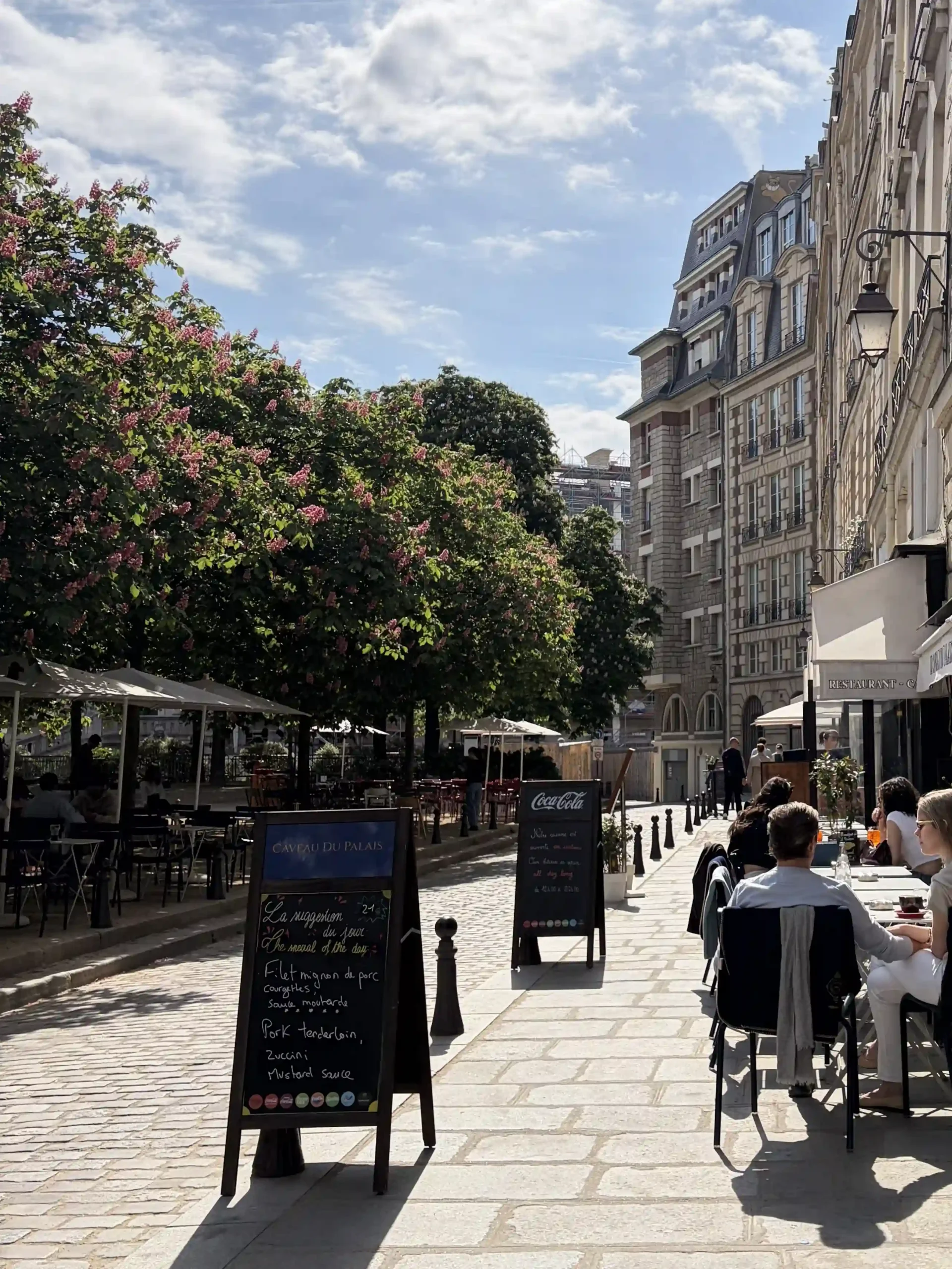 View of a cobblestone street in Paris. People sitting outside Caveau du Palais enjoing the sun.