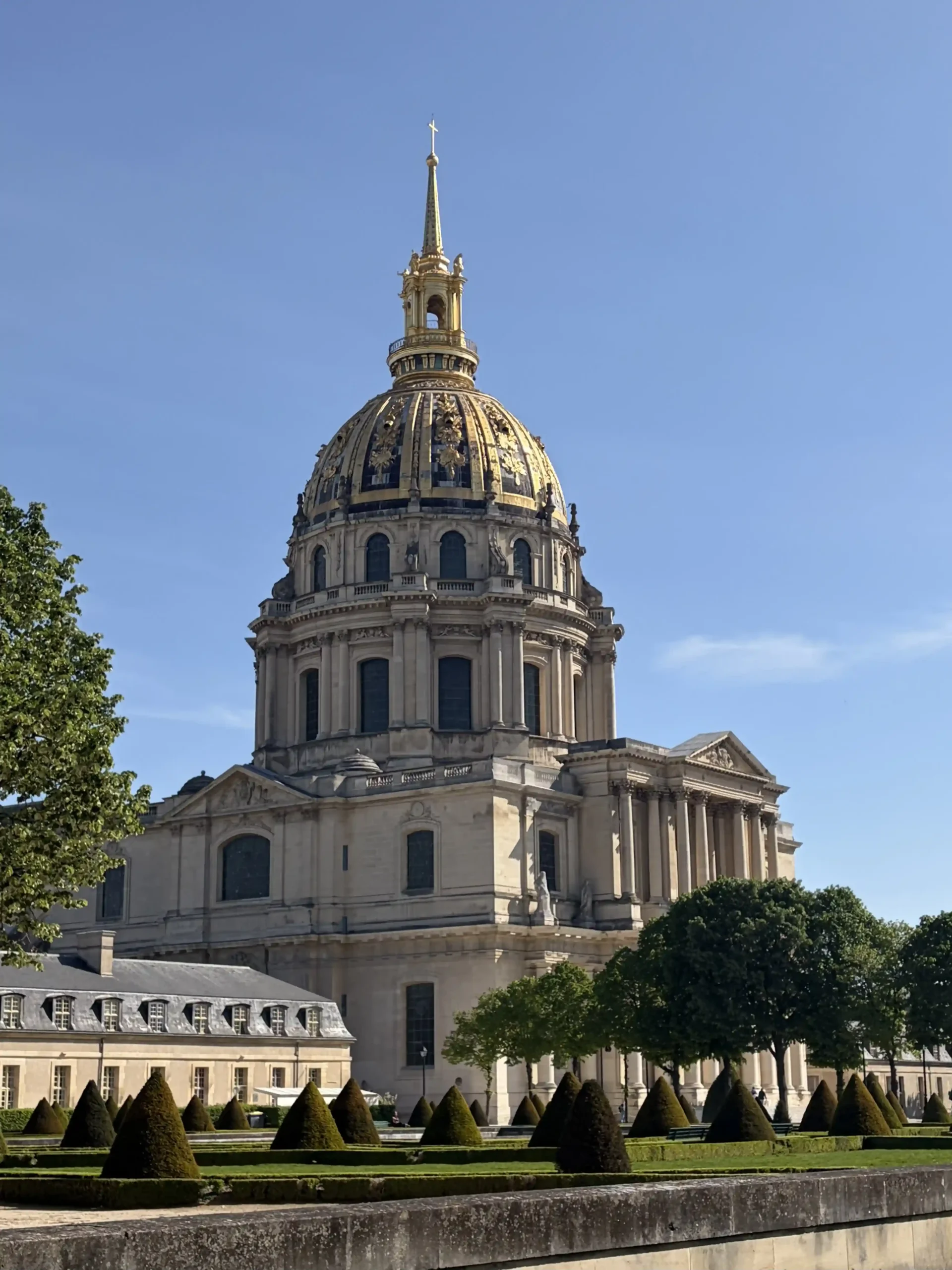 View of the Les Invalides monument in Paris. The building features a large stone structure topped with a golden dome and spire.