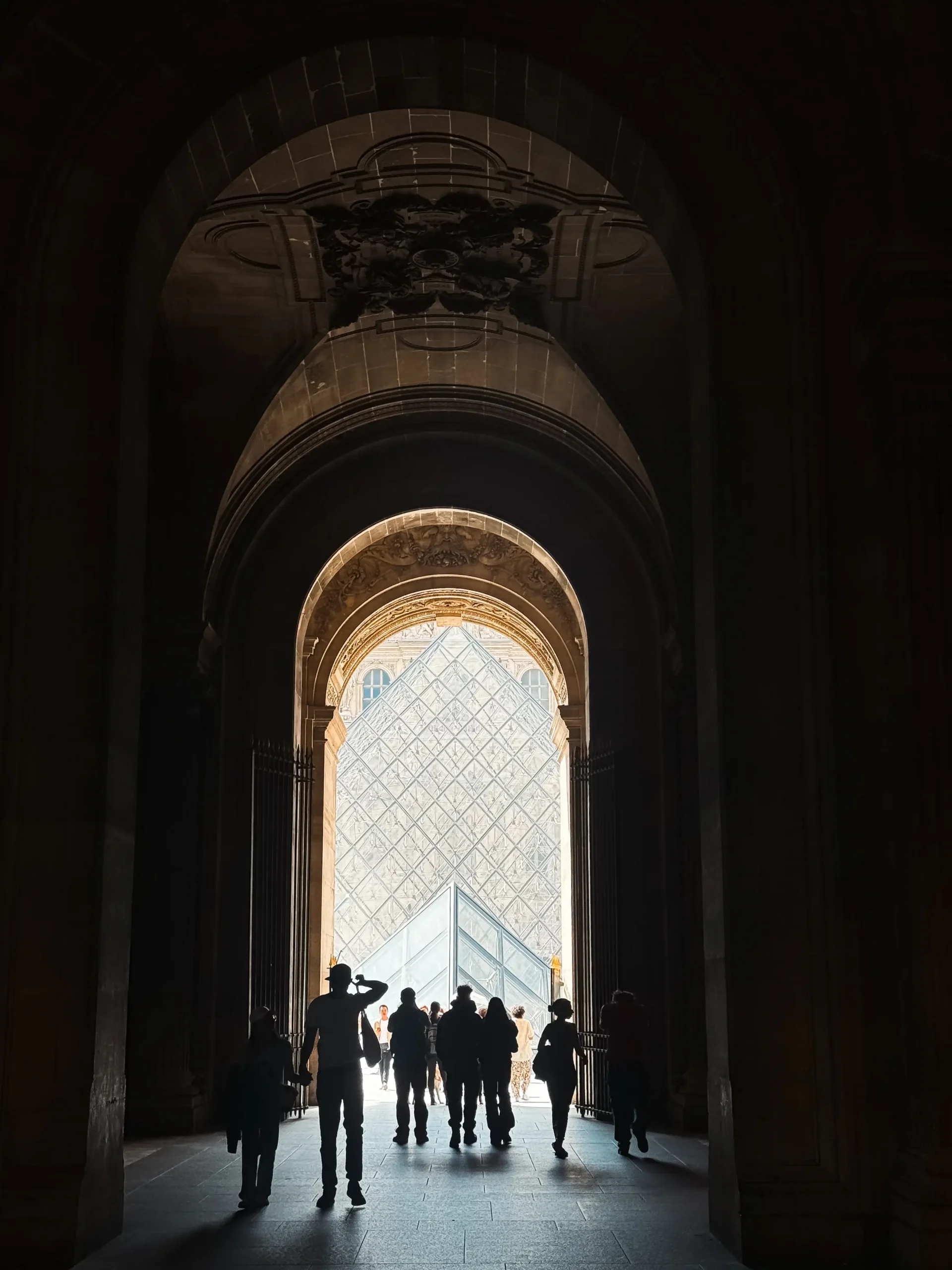 View through a dark, vaulted stone archway showing silhouetted pedestrians walking toward the bright glass Louvre Pyramid in Paris.