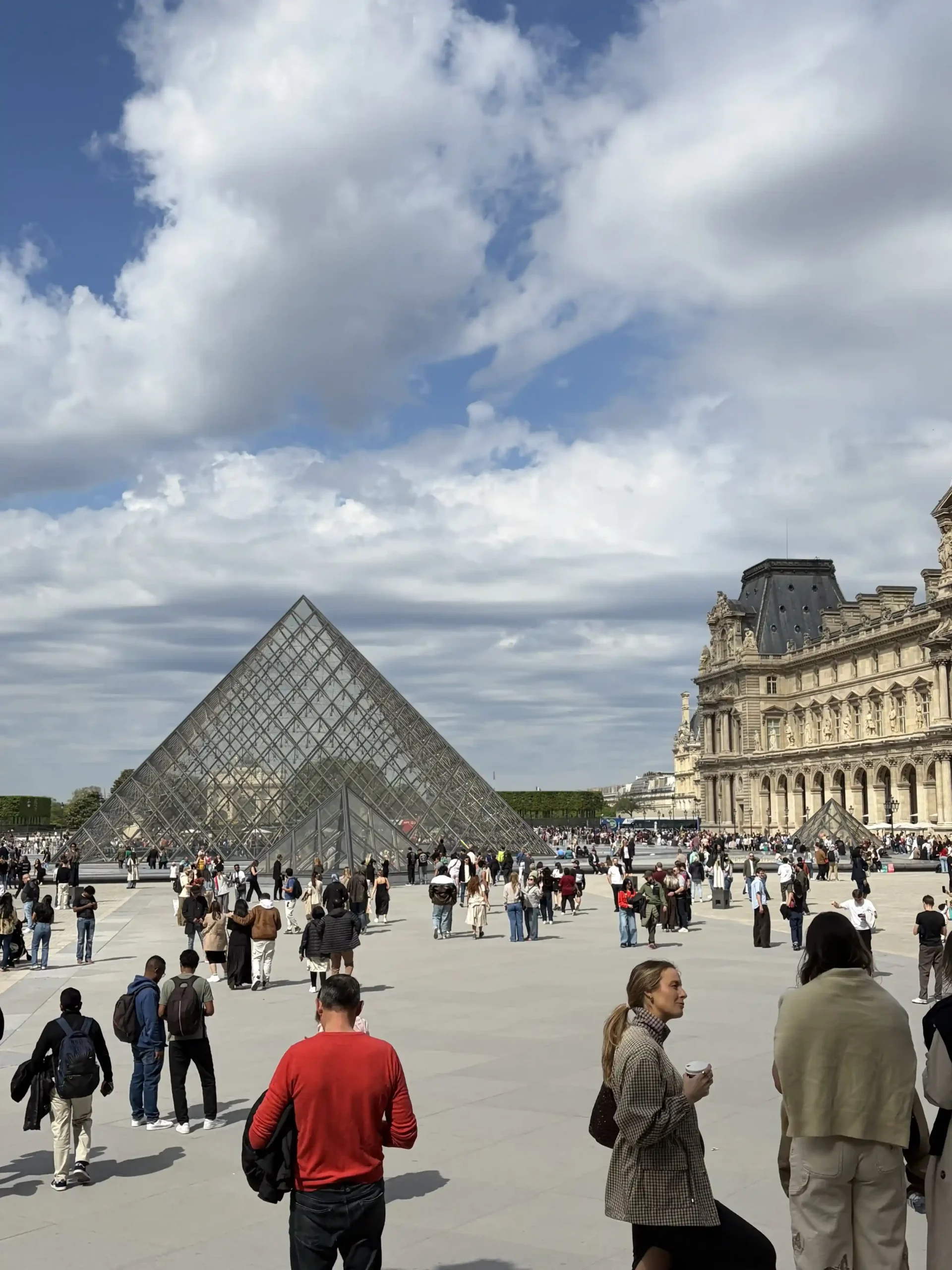 On the left sits the large glass and metal Louvre Pyramid, surrounded by smaller glass pyramids. To the right stands the ornate, classic architecture of the museum\'s palace wing. A large, diverse crowd of tourists walks across the expansive, light-colored stone plaza.