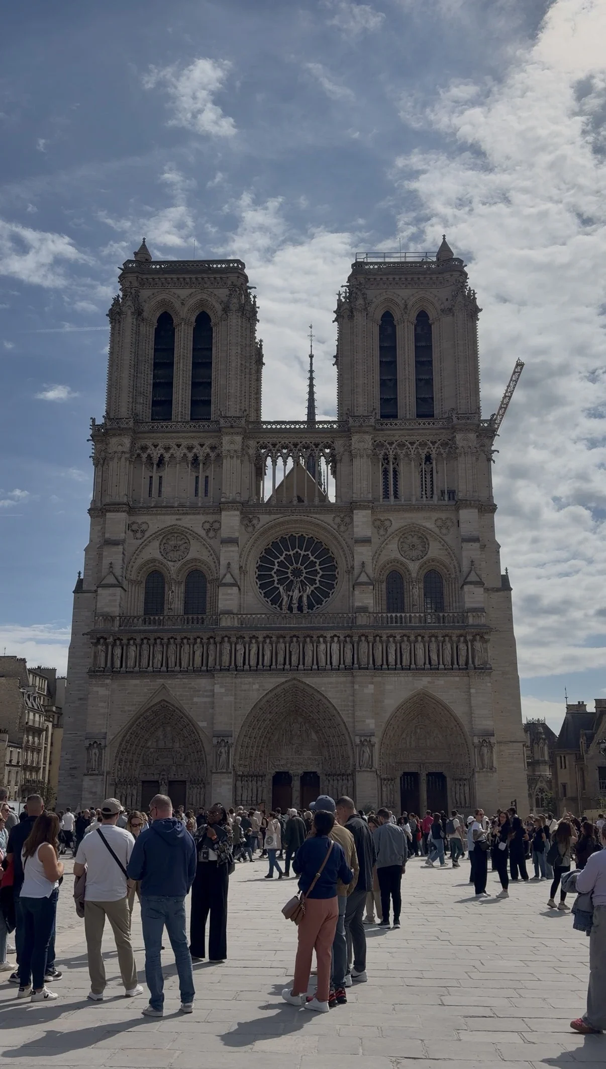 View of the historic stone Notre-Dame Cathedral features two massive square towers, a central rose window, and grand arched entryways. A large crowd of tourists walks across the open stone plaza in the foreground.