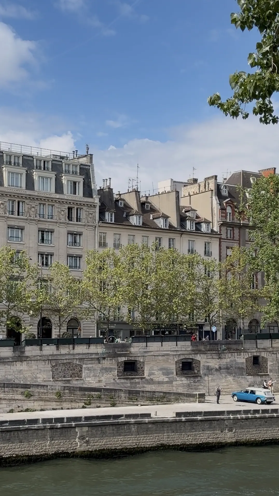 view of historic stone apartment buildings with mansard roofs. Green trees line the upper street level, while a lower stone embankment features a vintage blue station wagon parked near the water.