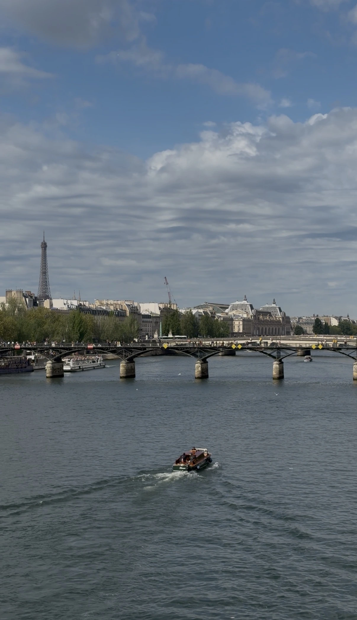 A small motorboat heading toward the multi-arched Pont des Arts bridge. In the distant background, the Parisian skyline is visible alongside the Eiffel Tower.