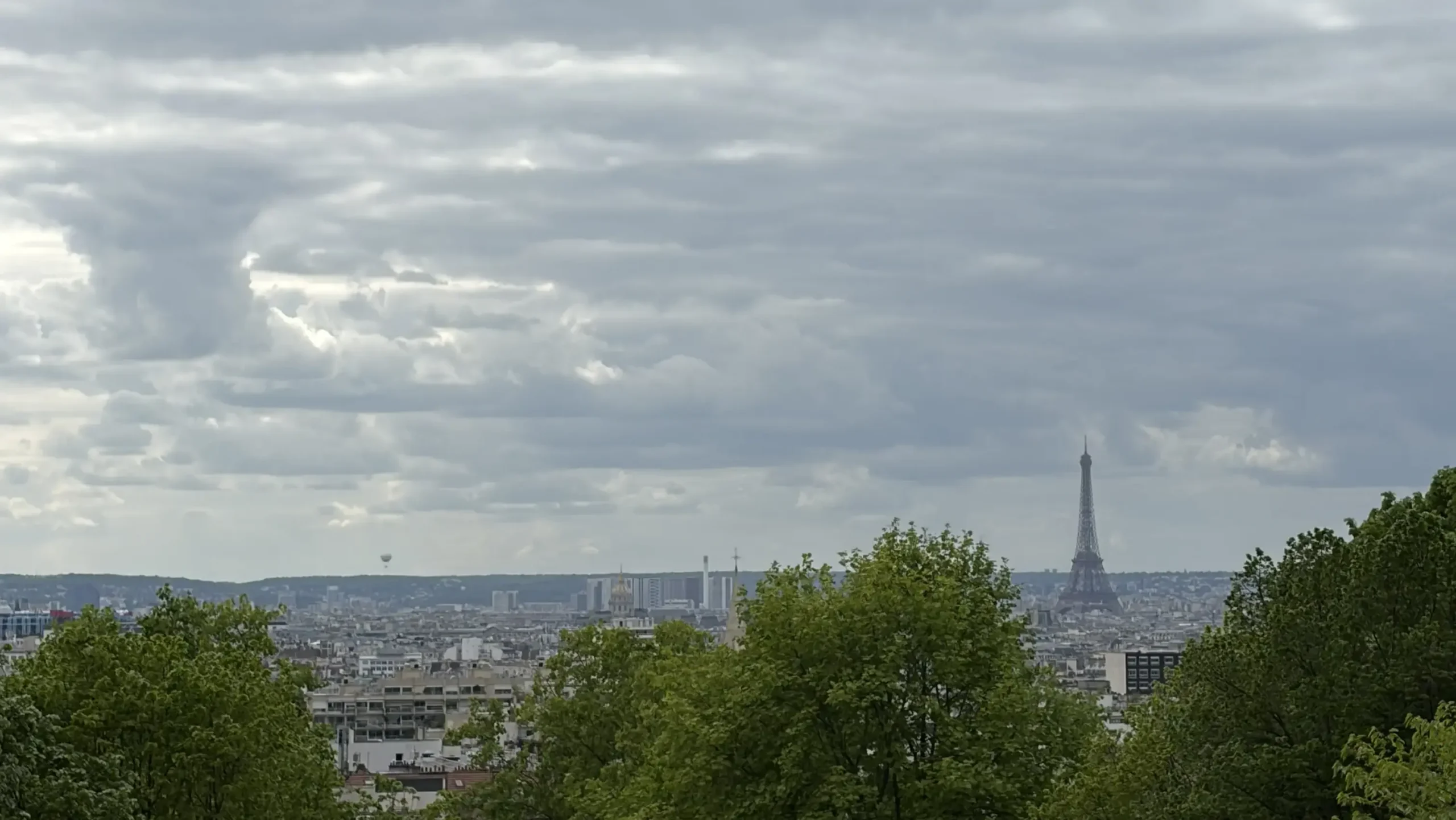 Wide shot of the Paris skyline viewed from an elevated vantage point. Thick, lush green tree canopies line the foreground, framing a distant view of urban buildings and the distinct silhouette of the Eiffel Tower.